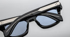 Close-up of Jacques Marie Mage Dealan 53 eyeglasses with thick black frames, blue-tinted lenses, and silver metallic detail on the arms, resting upside down on a light gray surface.