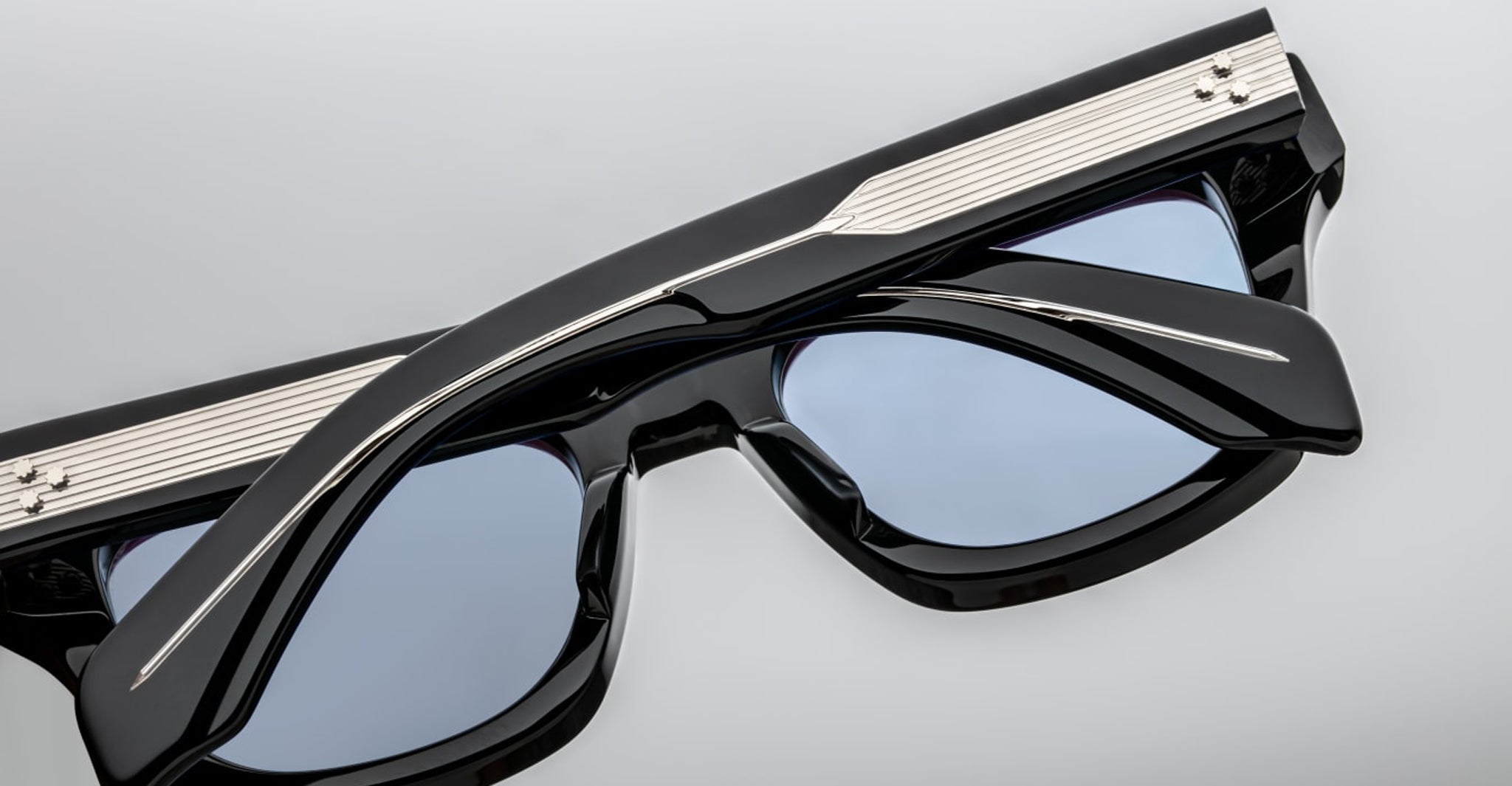 Close-up of Jacques Marie Mage Dealan 53 eyeglasses with thick black frames, blue-tinted lenses, and silver metallic detail on the arms, resting upside down on a light gray surface.