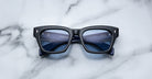 A pair of Jacques Marie Mage Dealan 53 rectangular black eyeglasses with blue-tinted lenses rests on a white marble surface, revealing printed text on the inside of the arms.