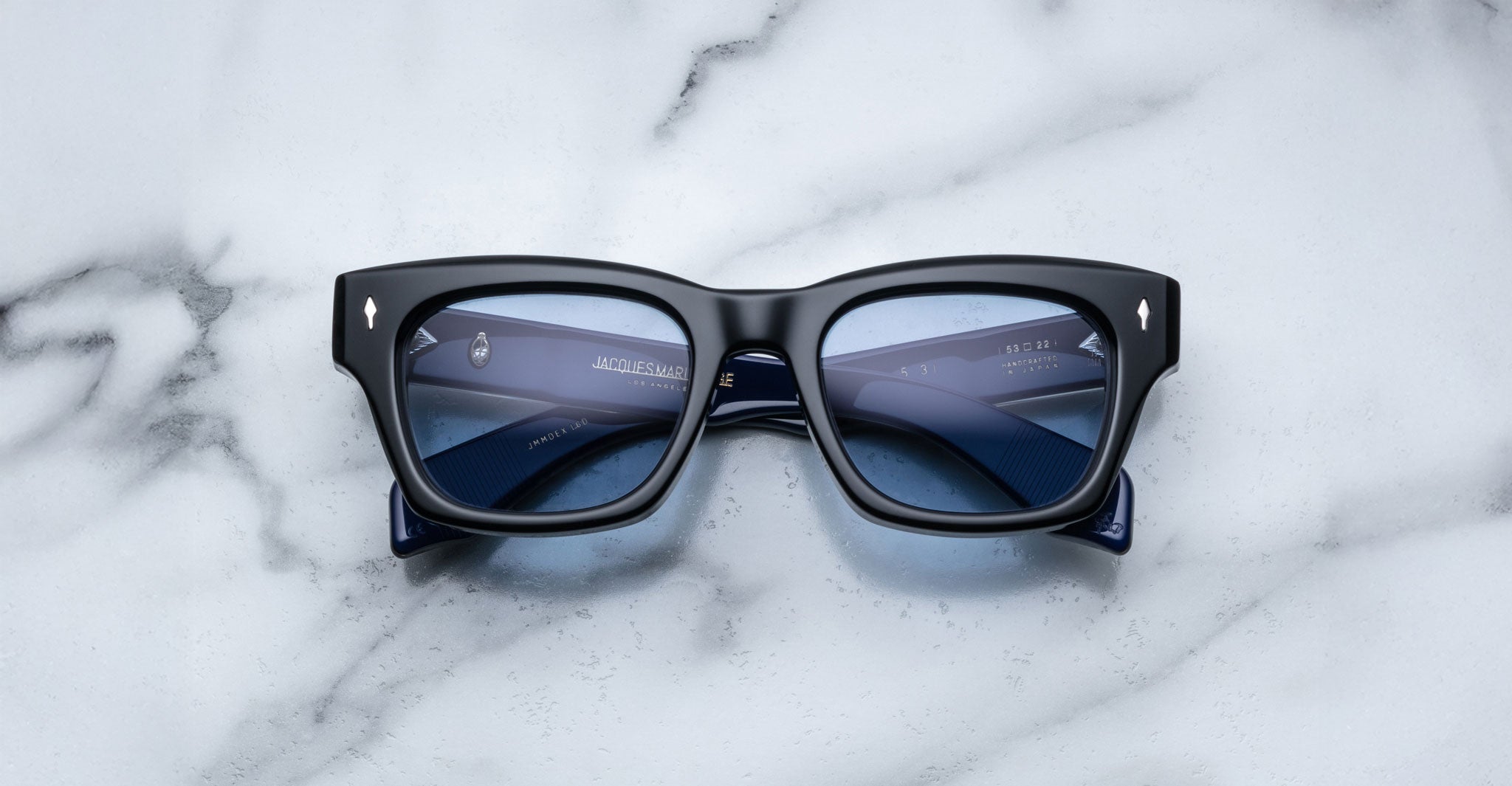 A pair of Jacques Marie Mage Dealan 53 rectangular black eyeglasses with blue-tinted lenses rests on a white marble surface, revealing printed text on the inside of the arms.