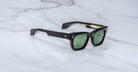 The Jacques Marie Mage Dealan 53 sunglasses, featuring a black rectangular frame, green lenses, and gold detailing on the arms, are placed on a white marble surface.