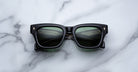 The Jacques Marie Mage Dealan 53 features black rectangular frames and dark green lenses, shown resting on a white marble surface with grey veining.