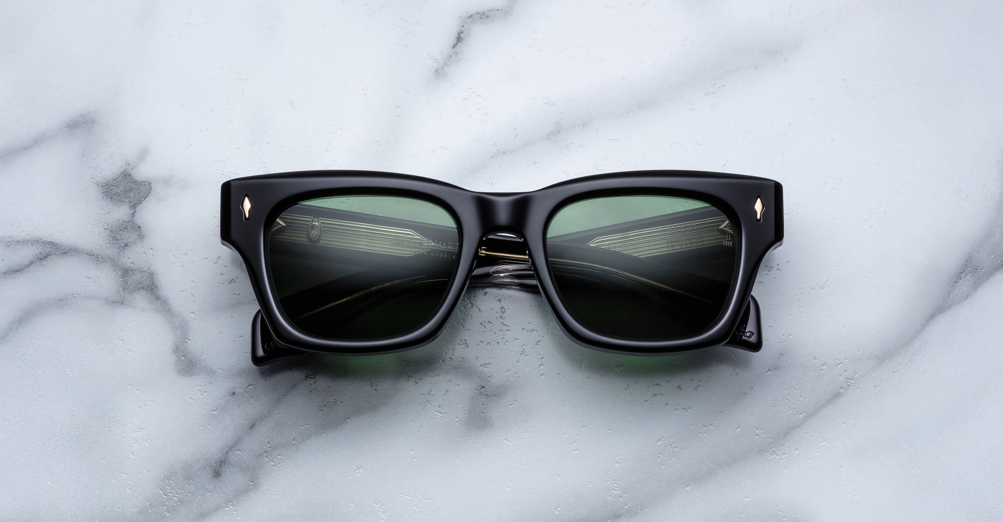 The Jacques Marie Mage Dealan 53 features black rectangular frames and dark green lenses, shown resting on a white marble surface with grey veining.
