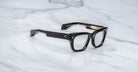 The Jacques Marie Mage Dealan 53, a pair of black rectangular eyeglasses with thick frames and clear lenses, is displayed on a white marble surface.