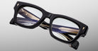 The Jacques Marie Mage Dealan 53 is a pair of black, thick-rimmed eyeglasses with decorative gold and black detailing on the inner arms, displayed against a light gray background.