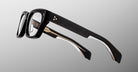 The Jacques Marie Mage Dealan 53 black rectangular eyeglasses feature thick frames, gold contacts by the left hinge, and a white accent along the inner arms, displayed from the side against a plain gray background.