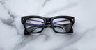 The Jacques Marie Mage Dealan 53, a pair of black rectangular eyeglasses with thick frames, rests on a white marble surface with its temples folded and visible through the lenses.