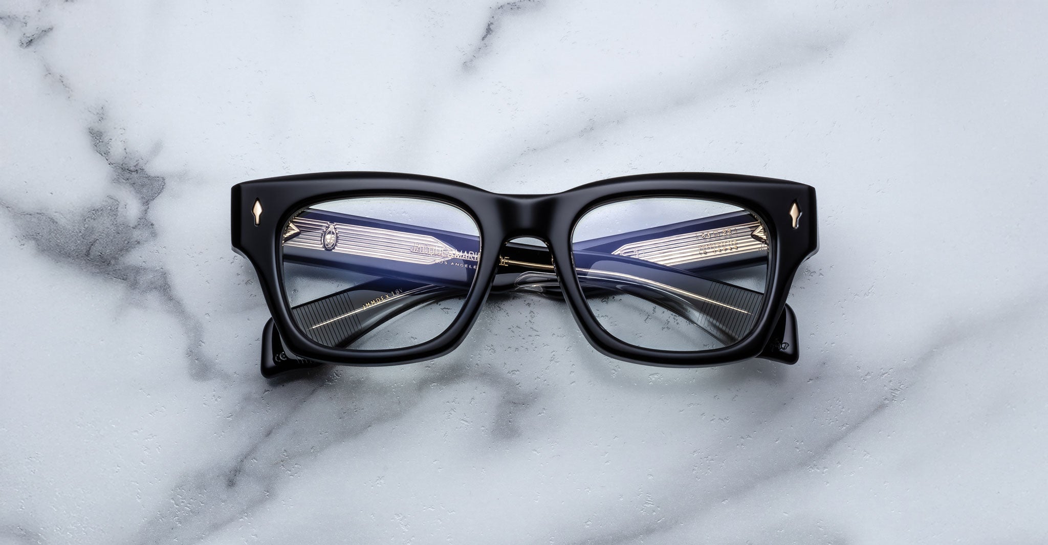 The Jacques Marie Mage Dealan 53, a pair of black rectangular eyeglasses with thick frames, rests on a white marble surface with its temples folded and visible through the lenses.