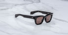 The Jacques Marie Mage Dealan 53 sunglasses, featuring black frames and brown tinted lenses, rest on a white marble surface. The temples show small silver dot accents and text appears on the inside of the right arm.