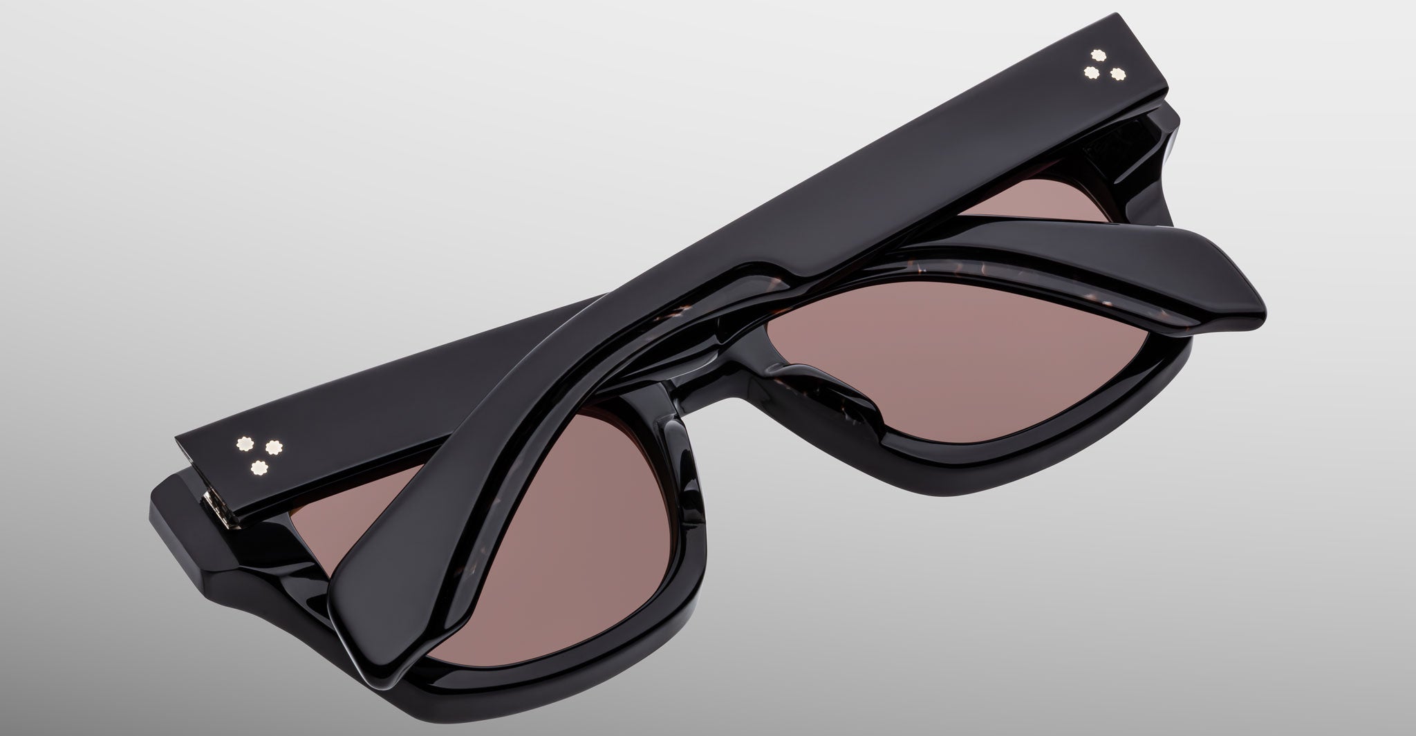 The Jacques Marie Mage Dealan 53 sunglasses, featuring black frames, brown tinted lenses, and decorative dots on the top corners, are shown folded closed against a light gray background.