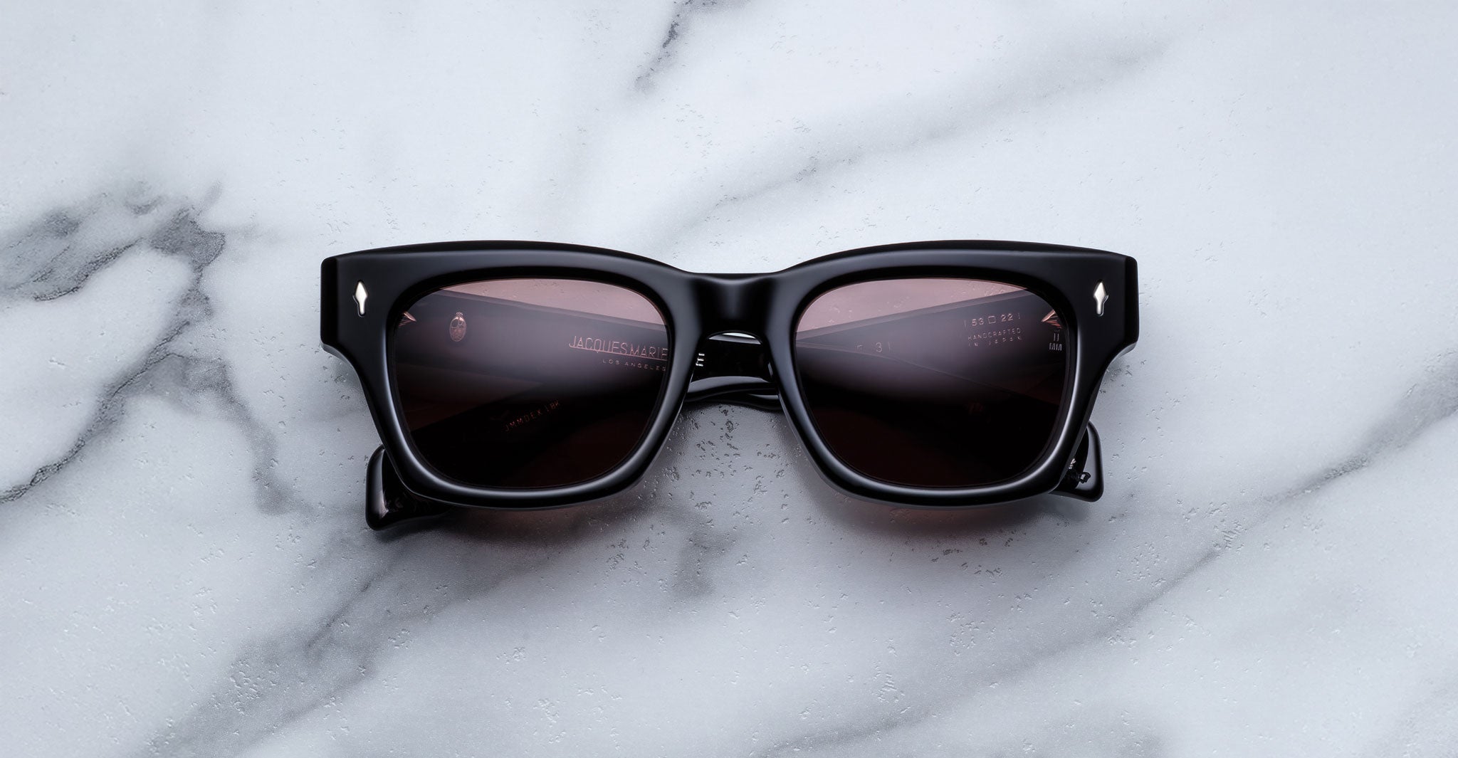 The Jacques Marie Mage Dealan 53, black rectangular sunglasses with dark lenses, are displayed on a white marble surface with grey veining.