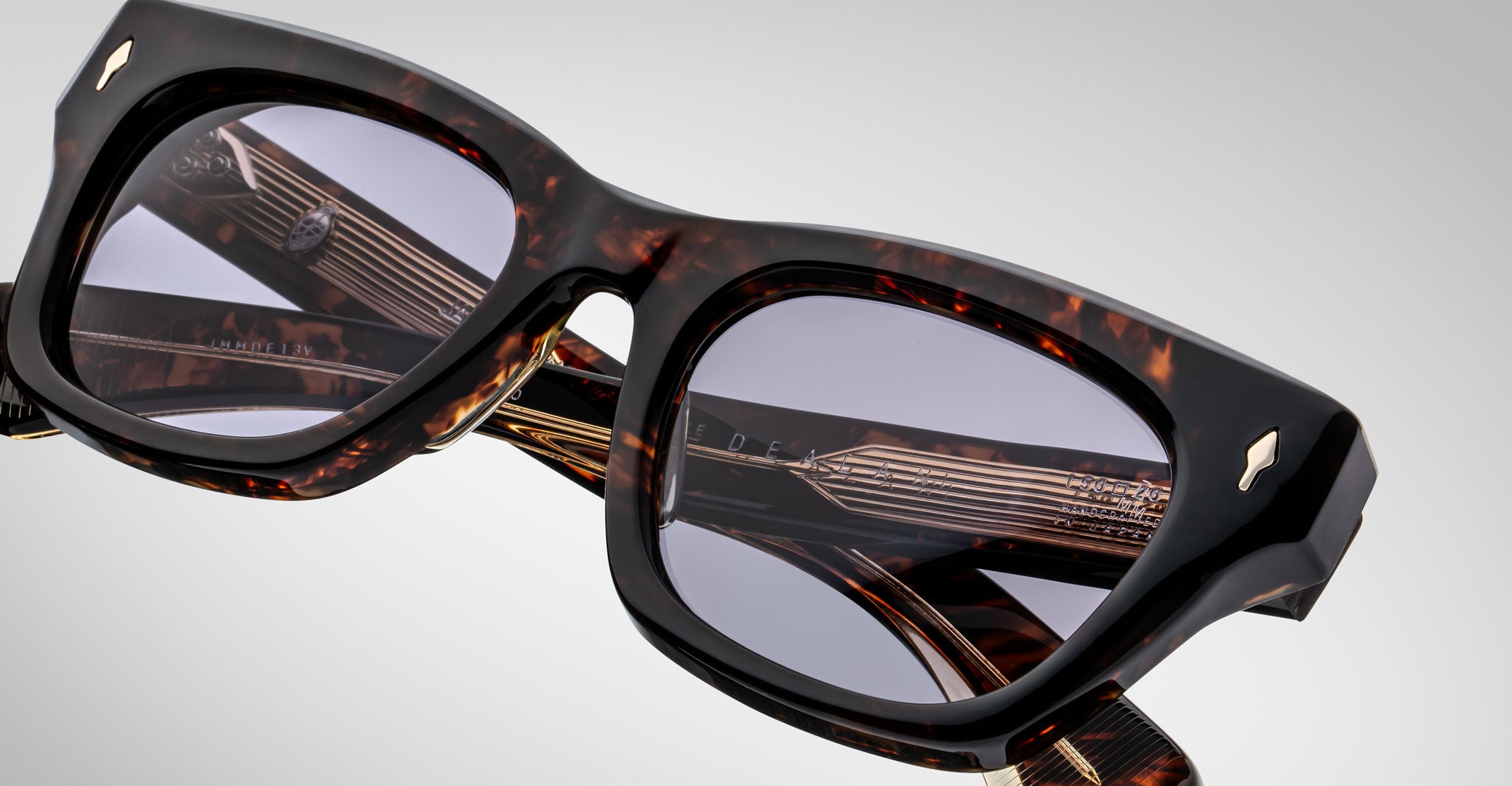 Close-up of Jacques Marie Mage Dealan eyeglasses featuring brown tortoiseshell thick frames, light gray tinted lenses, and metallic temple accents, highlighting texture against a gradient background.