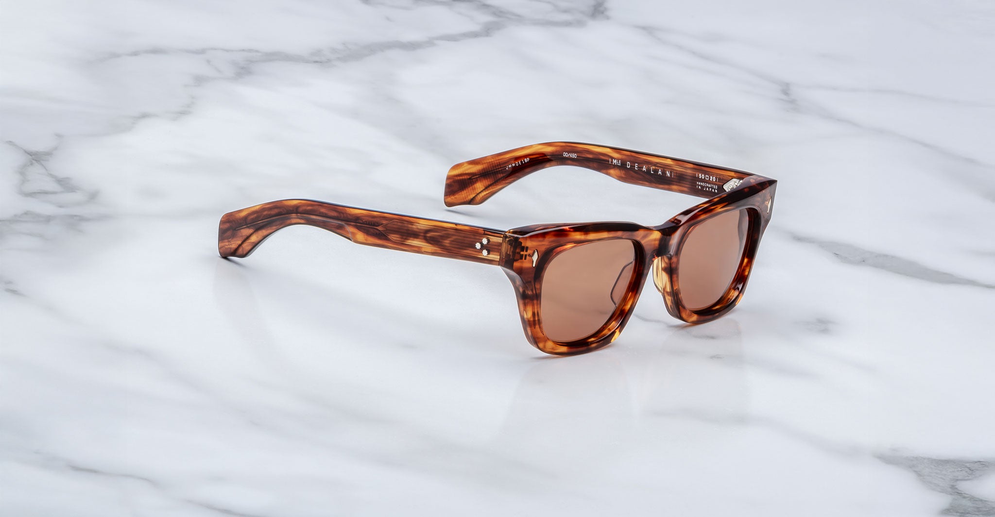 A pair of Jacques Marie Mage Dealan brown tortoiseshell sunglasses with rectangular lenses and thick frames rests on a white marble surface.