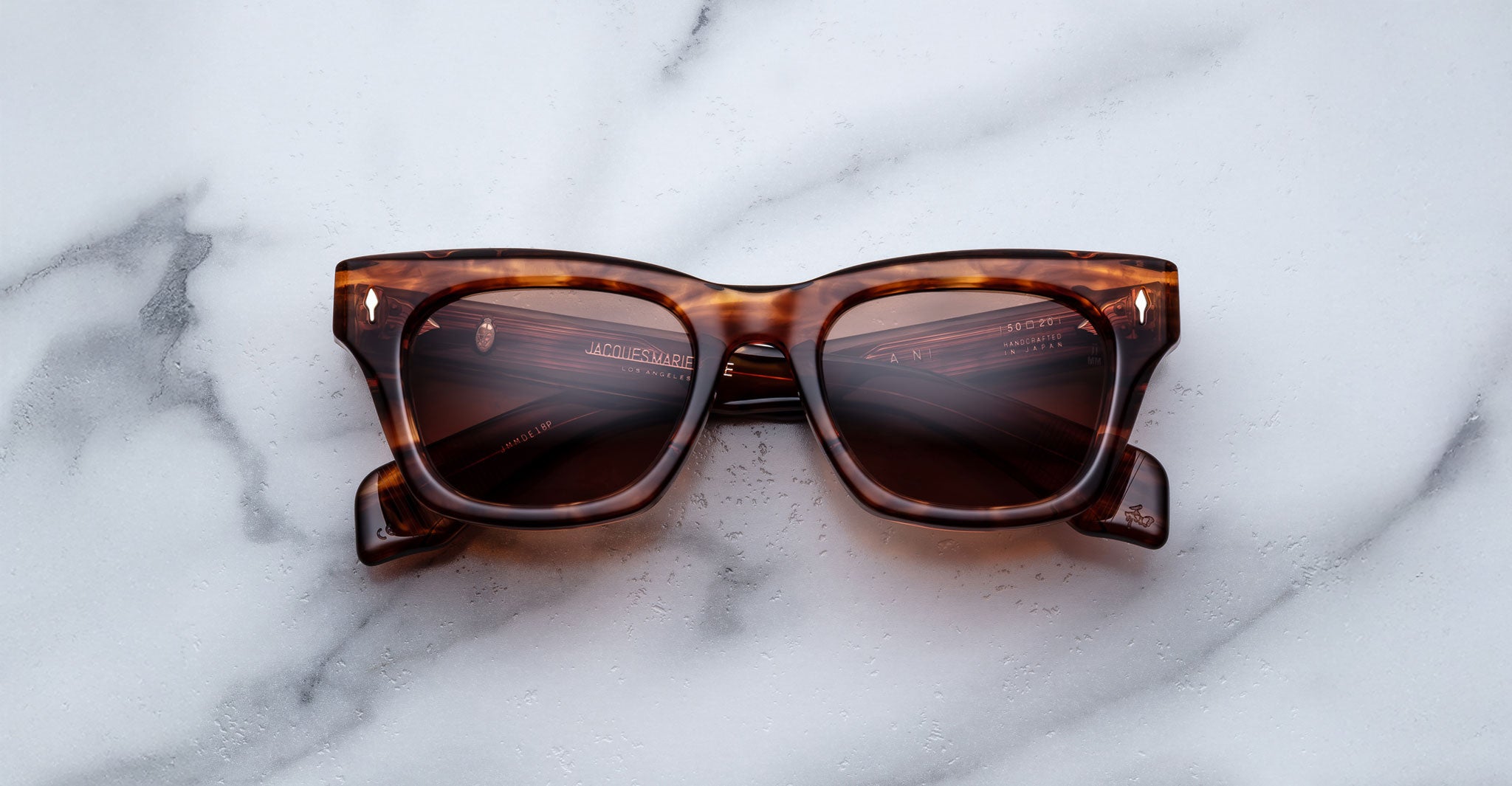 A pair of Jacques Marie Mage Dealan rectangular tortoiseshell sunglasses with dark lenses rests on a white marble surface.