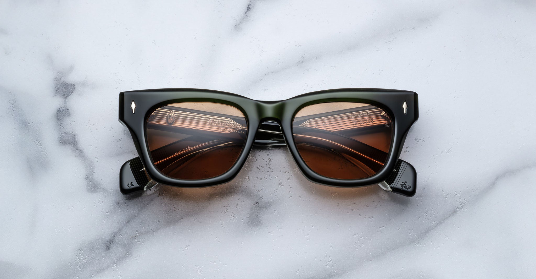 The Jacques Marie Mage Dealan sunglasses, featuring a black rectangular frame and brown tinted lenses, rest on a white marble surface. Reflections of curved lines and ceiling lights are visible on the lenses.