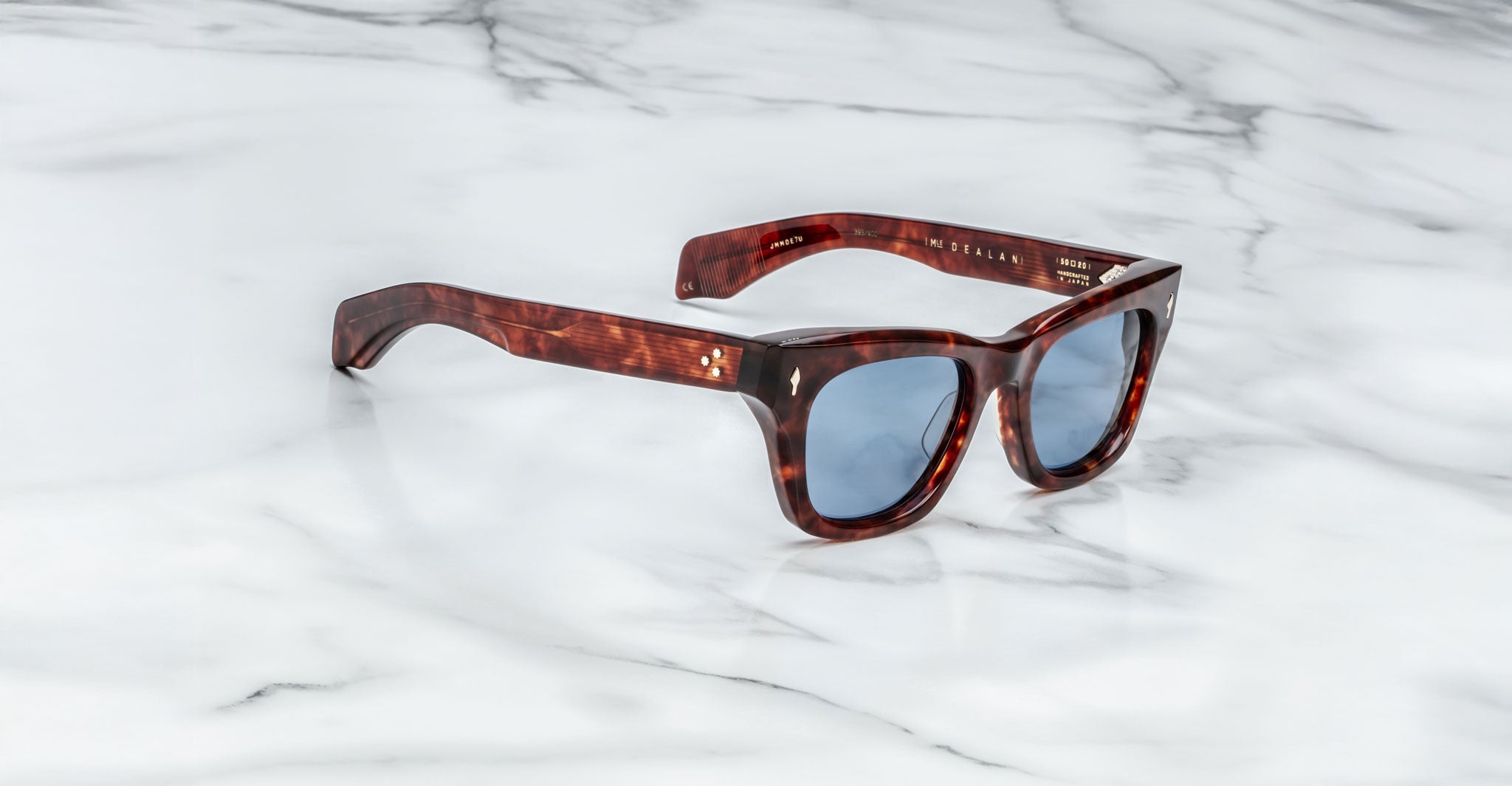 A pair of Jacques Marie Mage Dealan tortoiseshell sunglasses with blue-tinted lenses rests on a white marble surface, featuring gold text and small gold details near the hinges on the arms.