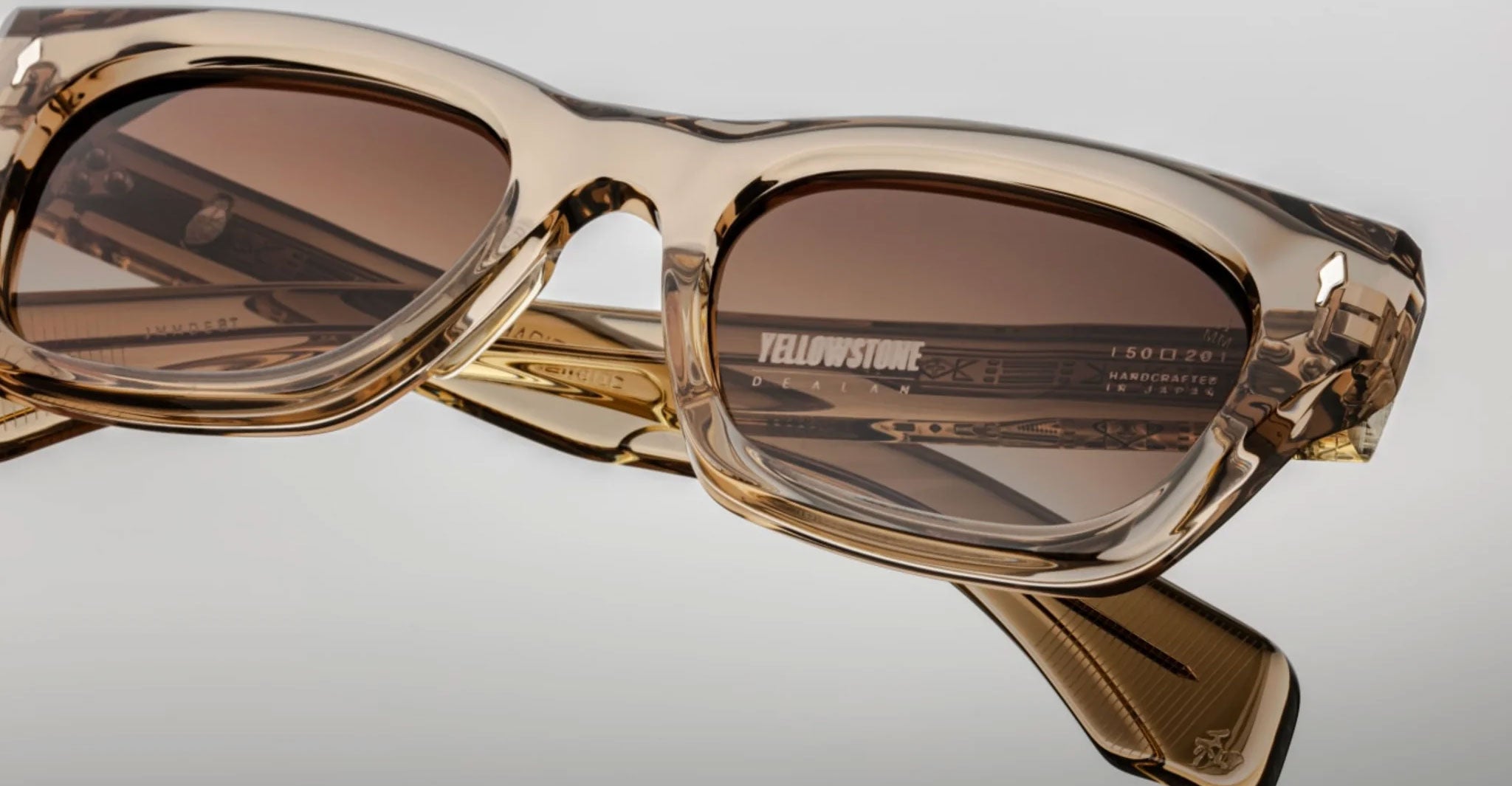 Close-up of Jacques Marie Mage Dealan translucent brown rectangular eyeglasses with dark tinted lenses, featuring “YELLOWSTONE” on one inner arm against a plain light background.