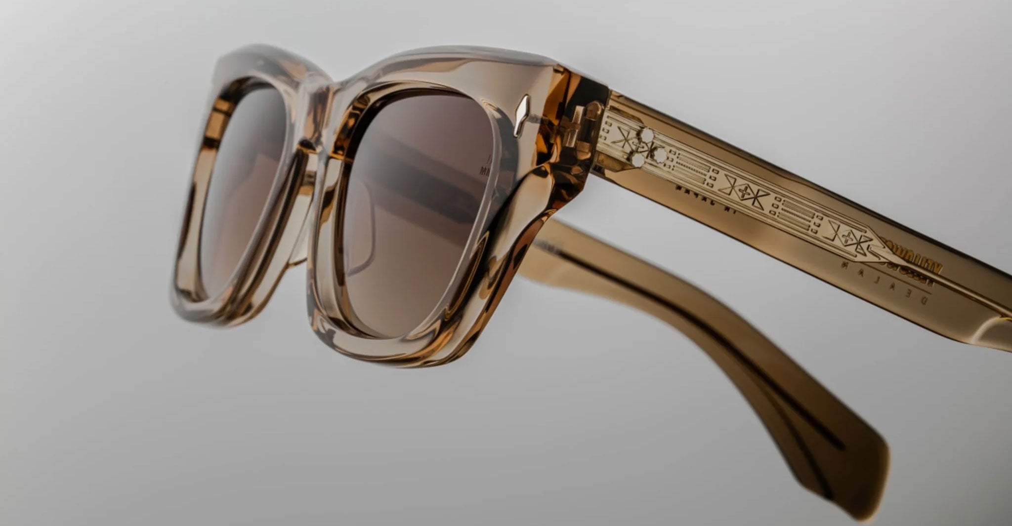 A close-up of Jacques Marie Mage Dealan sunglasses shows translucent brown square lenses and engraved symbols on the arms, set against a plain light background.