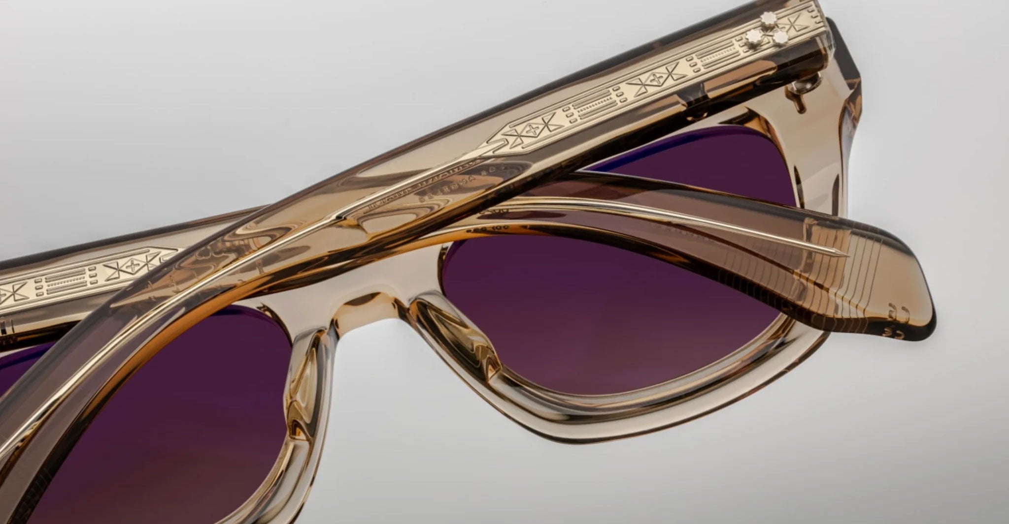 A close-up of two pairs of Jacques Marie Mage Dealan translucent brown sunglasses with purple lenses and etched geometric patterns on the arms, set against a plain light gray background.