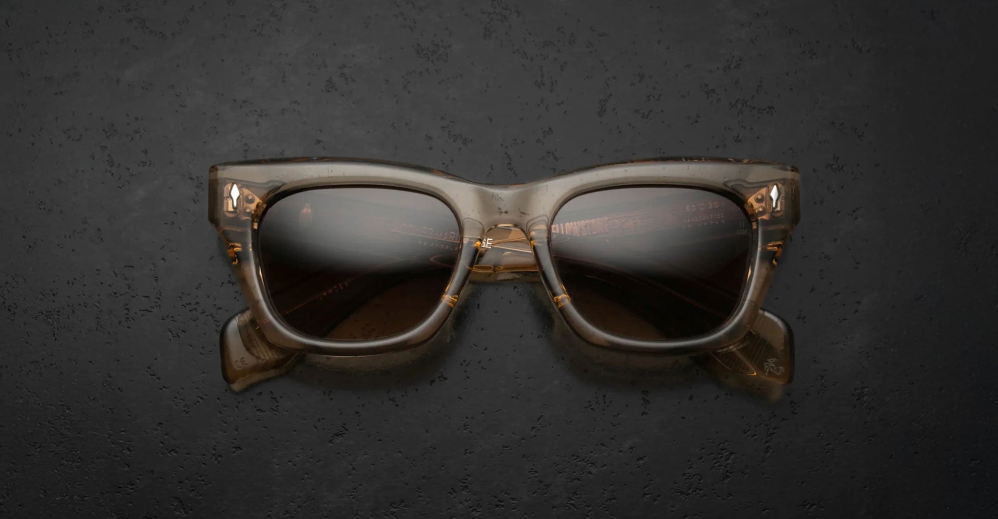 The Jacques Marie Mage Dealan sunglasses, featuring brown translucent square frames and dark lenses, are displayed from the front on a textured dark surface.