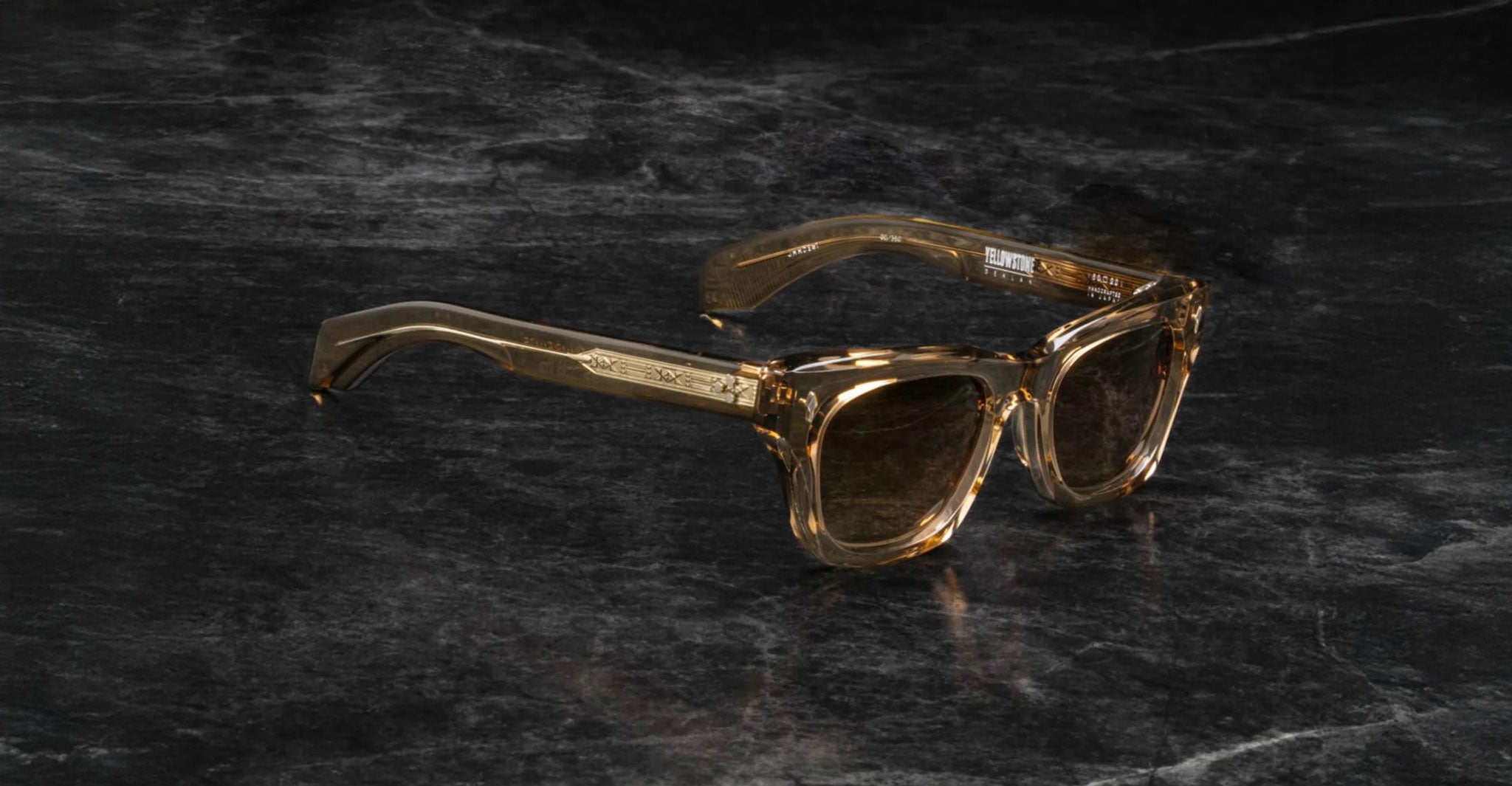 The Jacques Marie Mage Dealan features translucent brown square frames with decorative temple detailing, showcased on a dark marble surface.