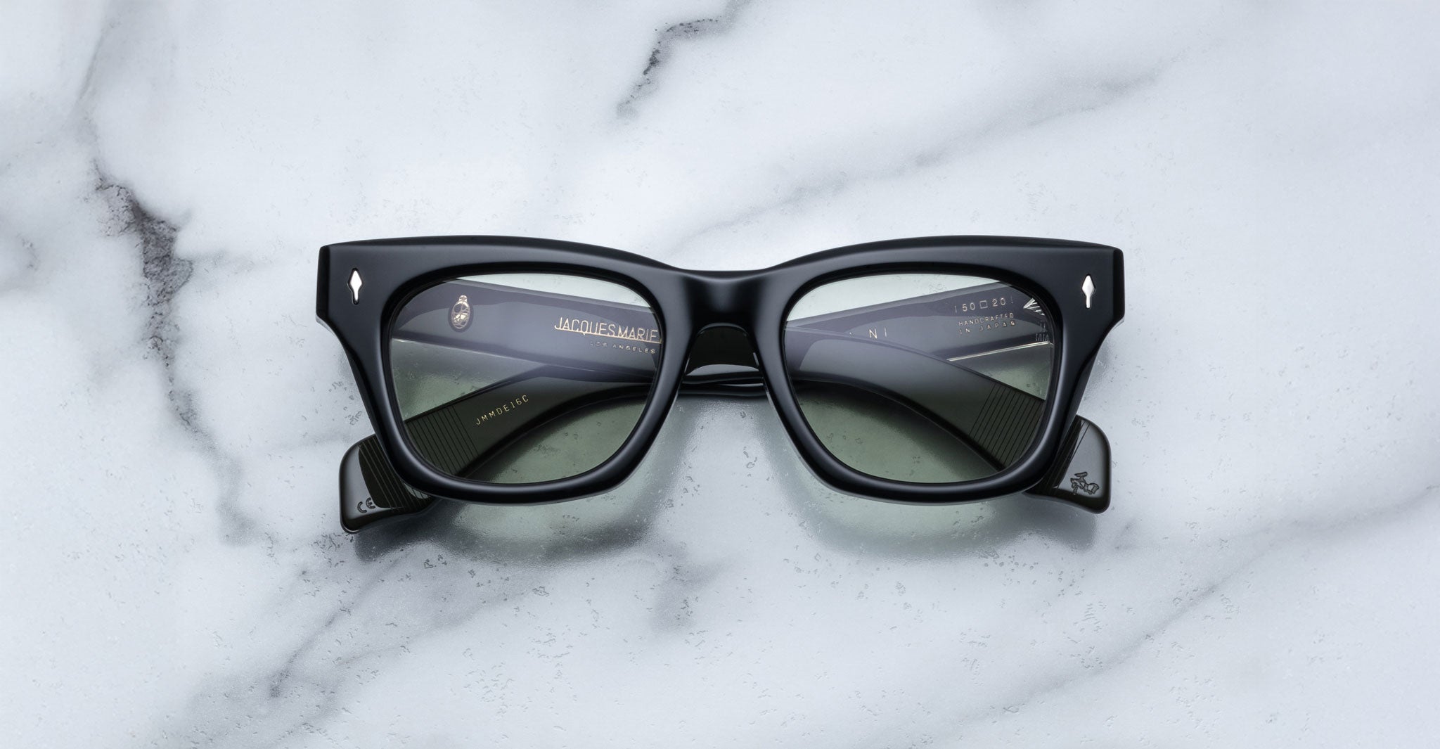 Jacques Marie Mage Dealan black rectangular sunglasses with dark tinted lenses rest on a white marble surface, showing gold brand markings and details on the inside arms.