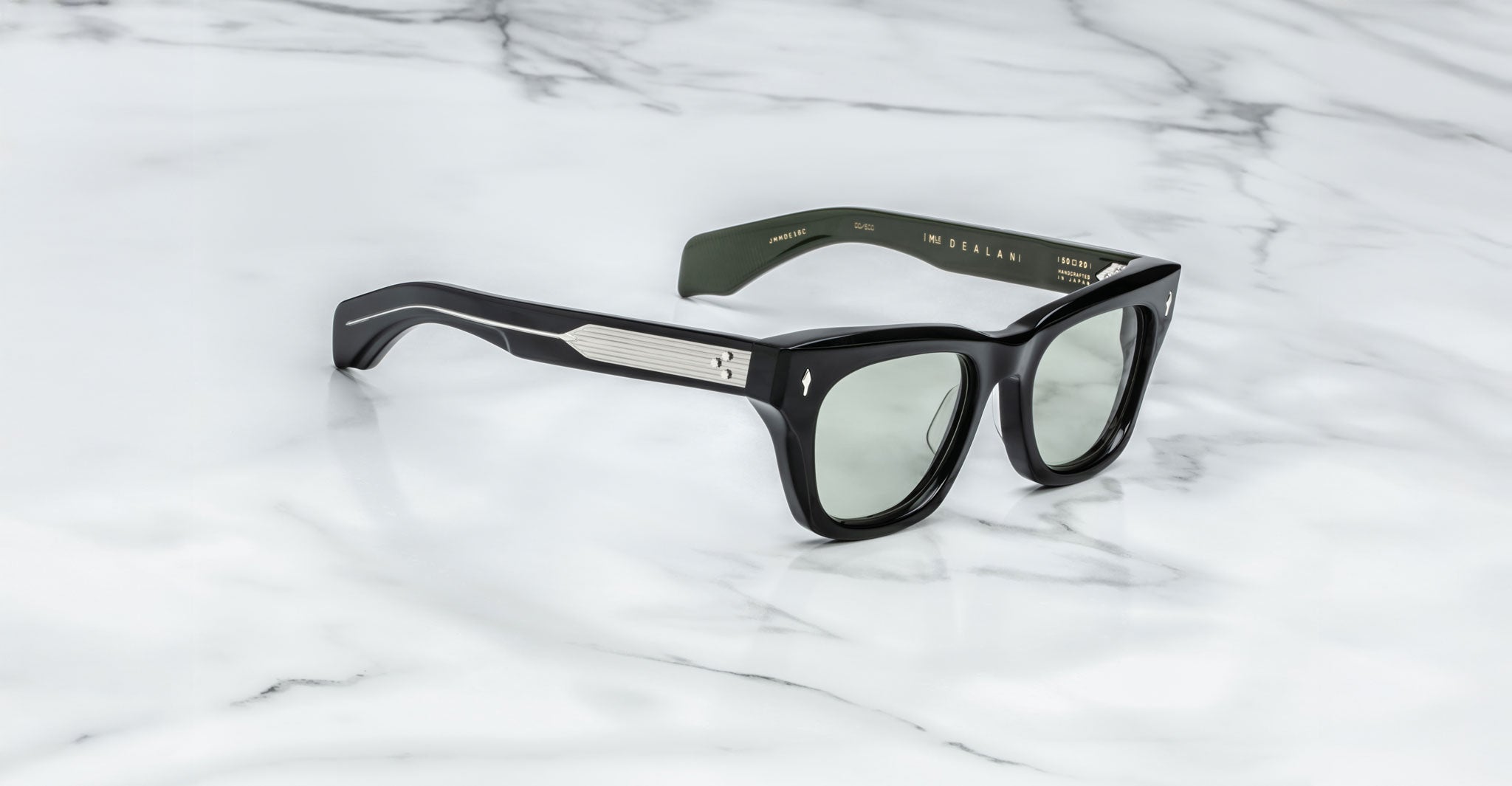 A pair of Jacques Marie Mage Dealan black rectangular sunglasses with silver metal accents on the arms, shown on a white marble surface. Gold text detailing the brand and model appears inside the arms.