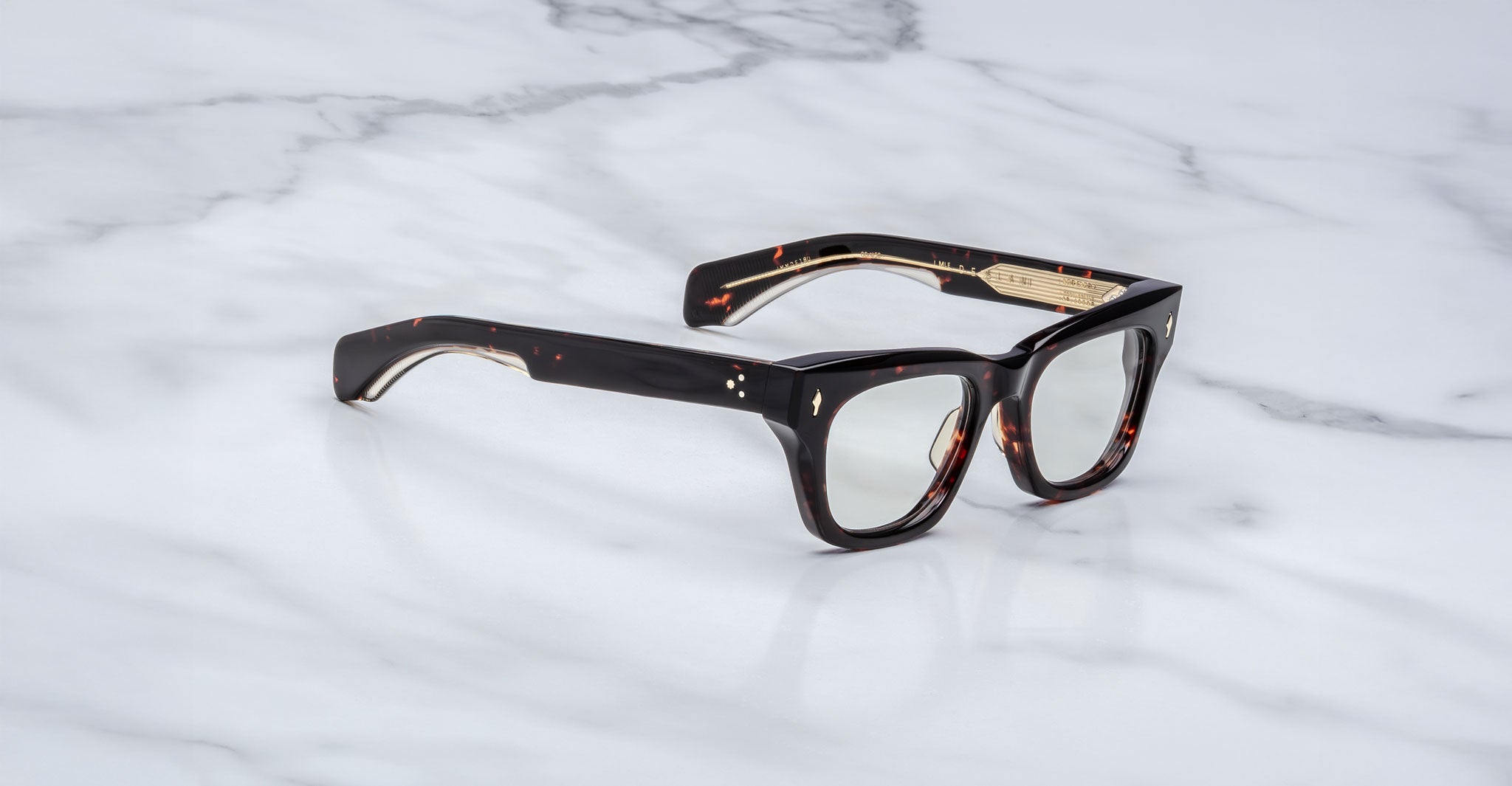 A pair of Jacques Marie Mage Dealan eyeglasses with dark tortoiseshell thick frames rests on a white marble surface.