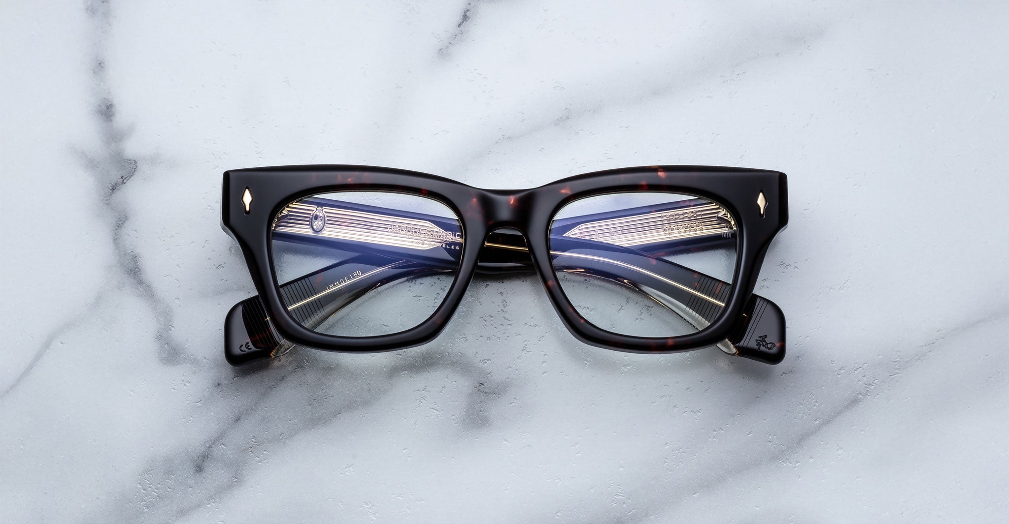 A pair of Jacques Marie Mage Dealan thick, rectangular black eyeglasses with a subtle tortoiseshell pattern rests on a white marble surface, their lenses reflecting lines of indoor lighting.