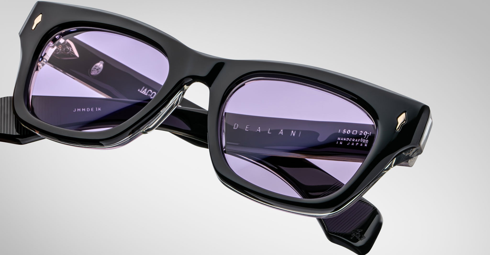 Close-up of Jacques Marie Mage Dealan black rectangular eyeglass frames with purple-tinted lenses. The inside temple arm shows the brand and manufacturing details against a plain, light background.