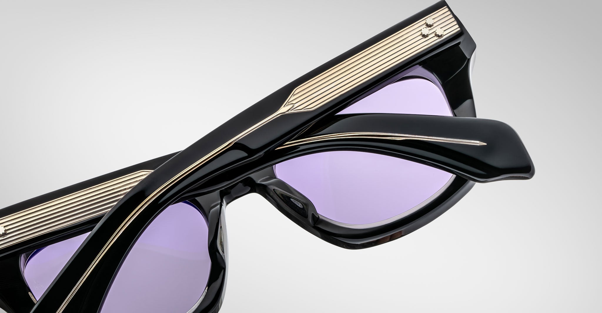 Close-up of folded Jacques Marie Mage Dealan sunglasses featuring black frames, gold detailing on the arms, and light purple-tinted lenses, set against a plain light gray background.
