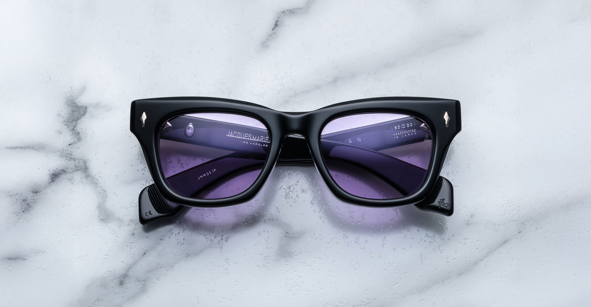 A pair of Jacques Marie Mage Dealan black rectangular sunglasses with purple-tinted lenses rests on a white marble surface, with branding and specifications visible on the inside arms.