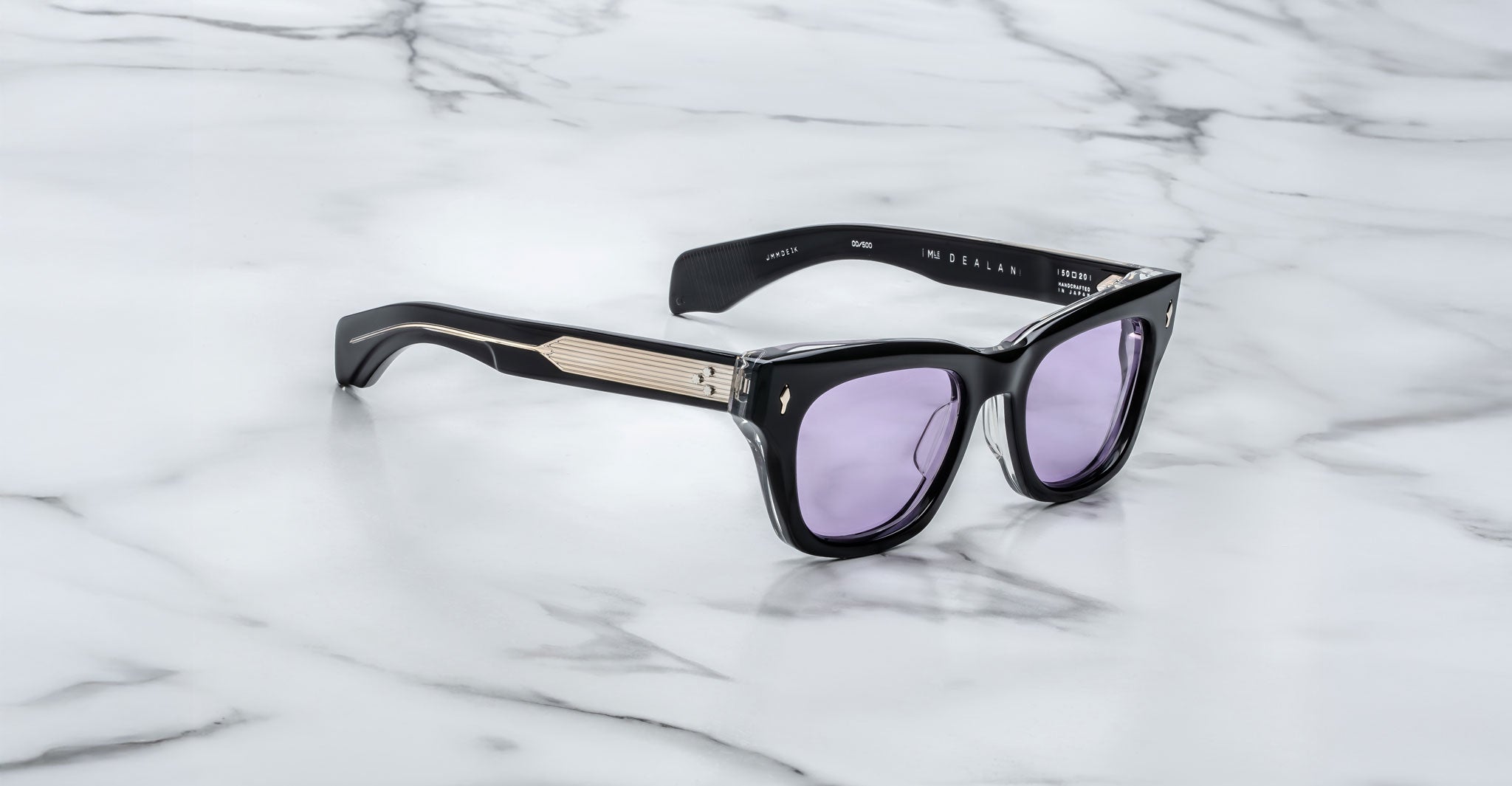 The Jacques Marie Mage Dealan sunglasses, featuring light purple-tinted lenses and metallic-accented frames, are displayed on a white marble surface. Printed text appears on the inner arms.