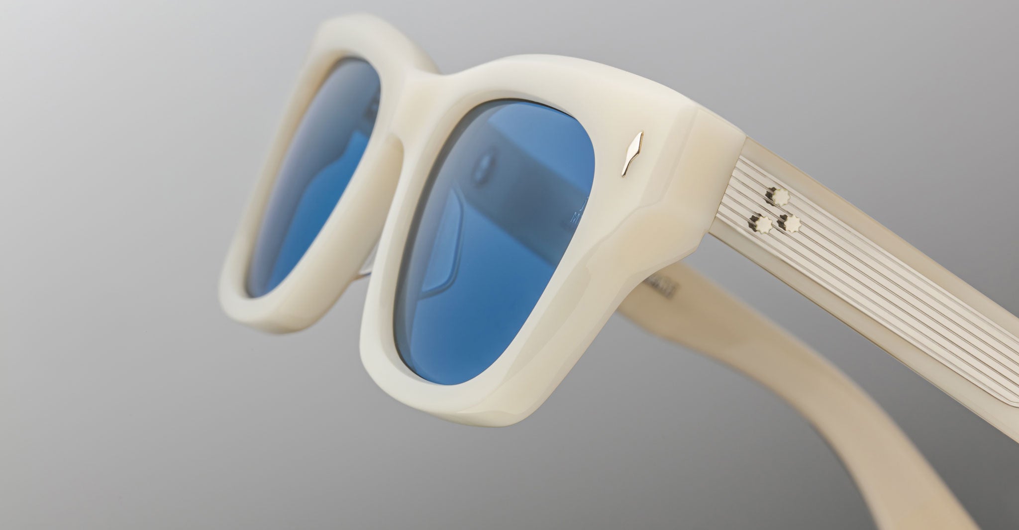 Close-up of Jacques Marie Mage Dealan sunglasses with thick off-white frames, blue-tinted lenses, metallic-accented temples, and a grooved design, set against a neutral gradient background.