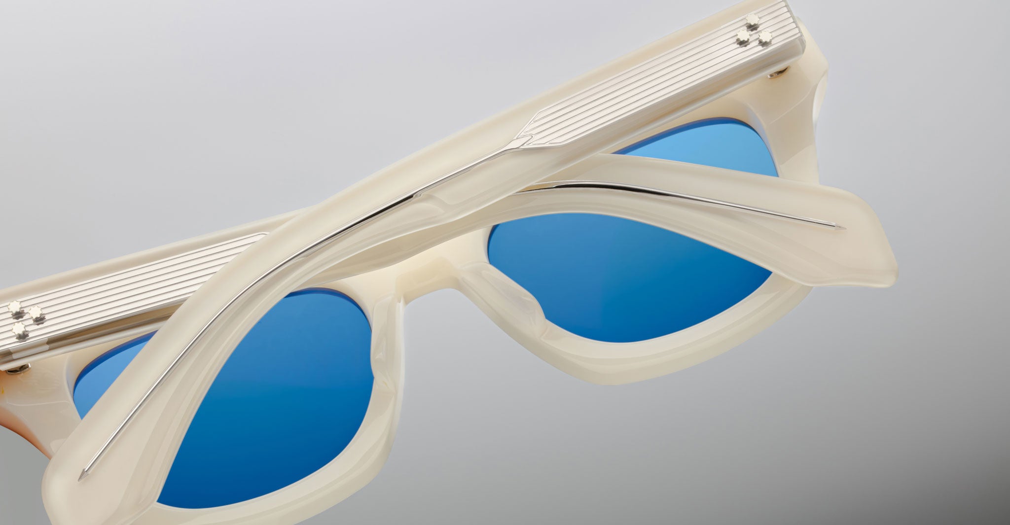 Close-up of Jacques Marie Mage Dealan sunglasses in beige with translucent frames and blue lenses, shown folded with arms crossed on a plain light gradient background.