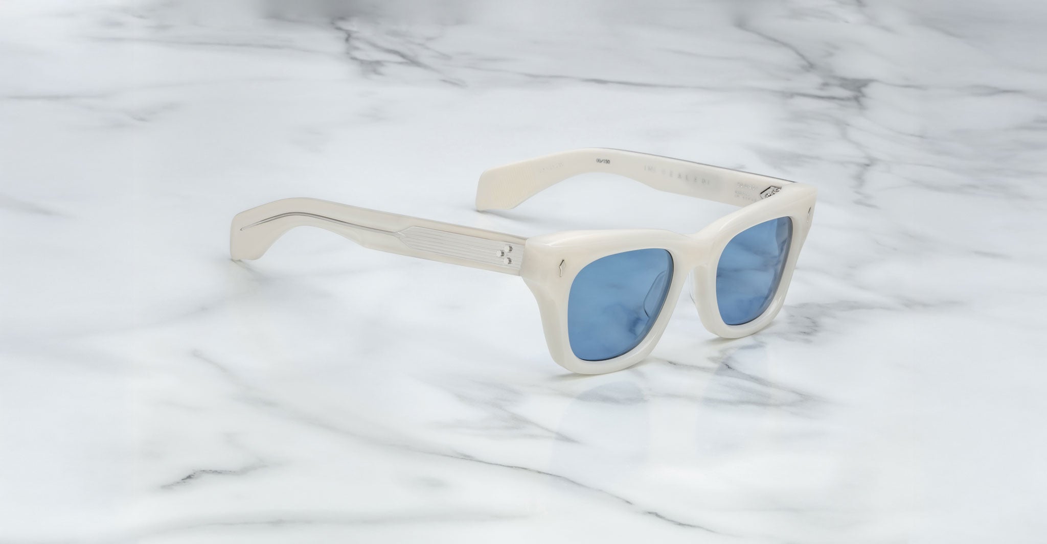 The Jacques Marie Mage Dealan sunglasses, featuring white frames and blue-tinted lenses, are angled on a white marble surface with grey veins, showcasing the left arm and front of the stylish frame.