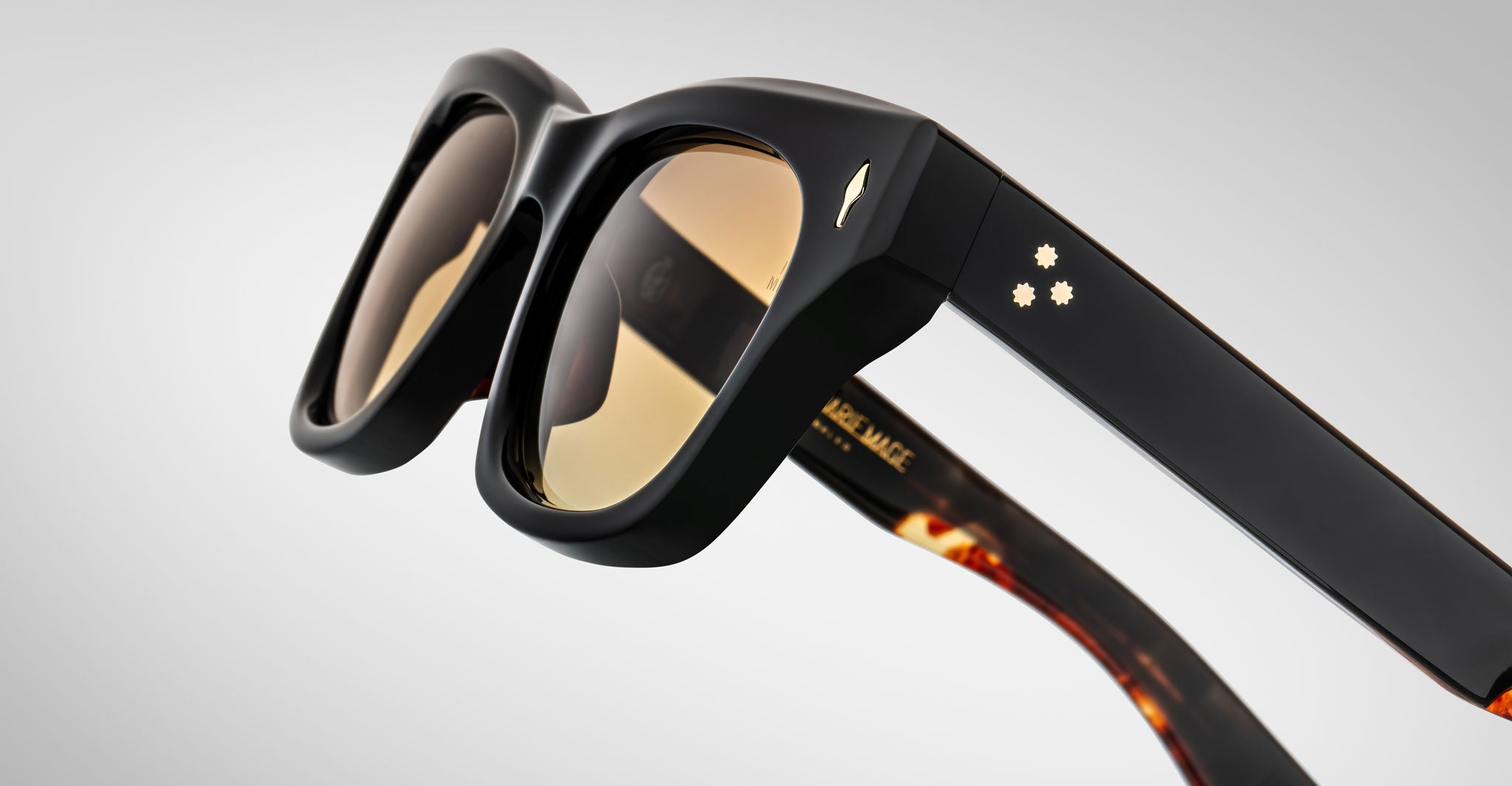 Close-up of Jacques Marie Mage Dealan sunglasses featuring thick black frames, amber-tinted lenses, and gold star accents on the temples against a softly lit plain background.