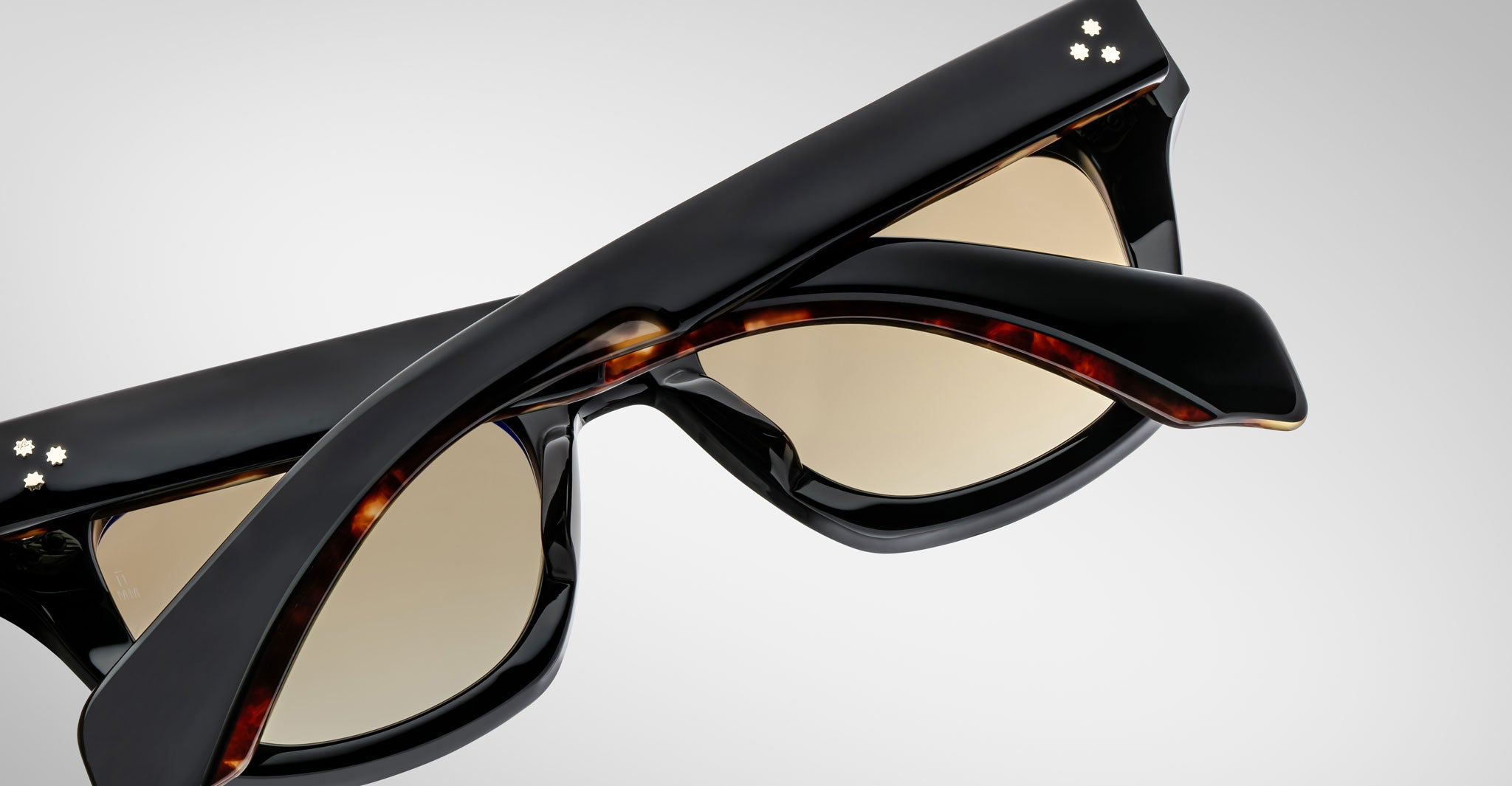 Close-up of folded Jacques Marie Mage Dealan sunglasses with black frames, tortoiseshell inner arms, and two small white dots on the hinges, set against a plain light background.
