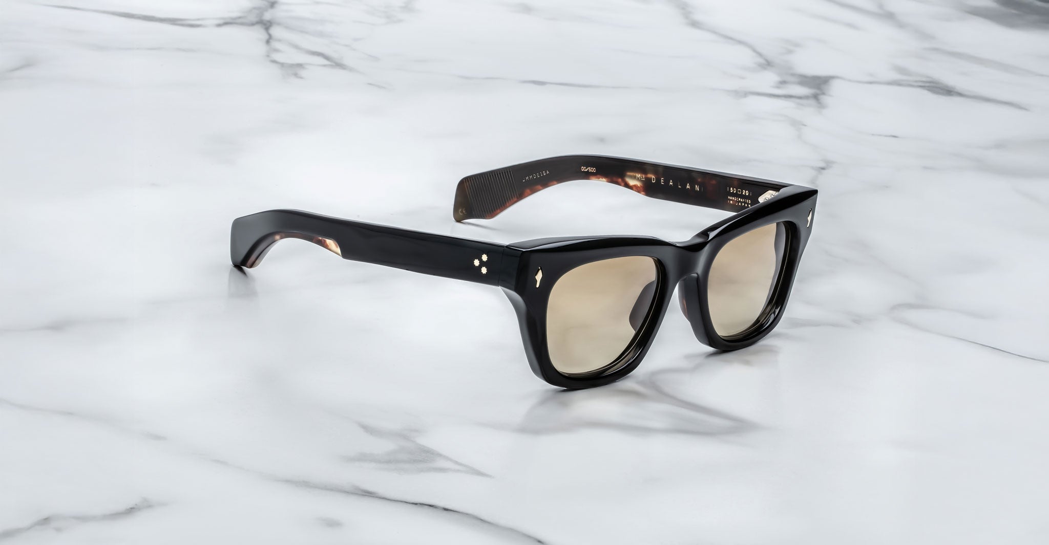 The Jacques Marie Mage Dealan sunglasses feature thick black rectangular frames, beige-tinted lenses, and small silver dots on the temples near the hinges, displayed on a white marble surface.