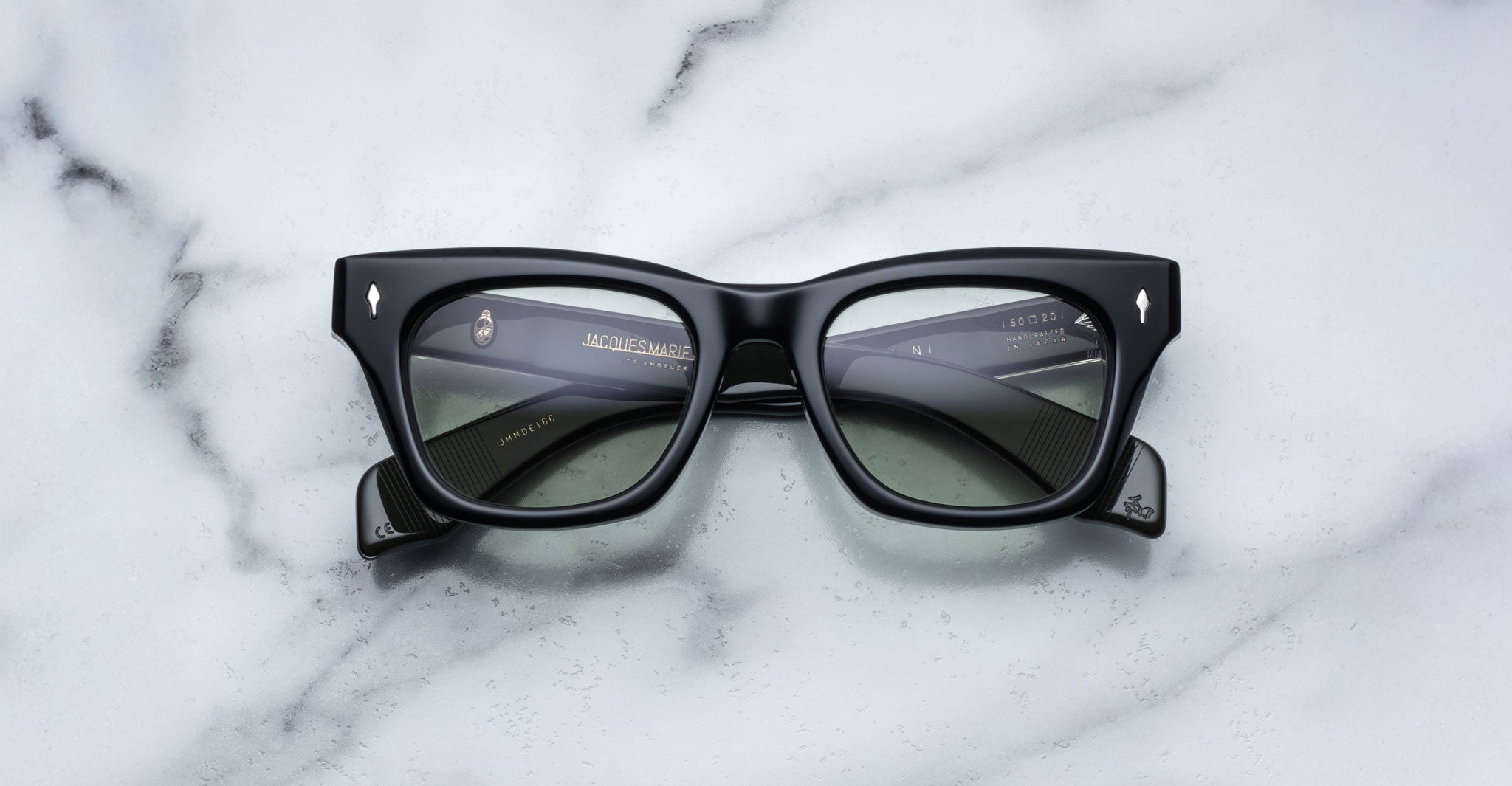 A pair of Jacques Marie Mage Dealan black rectangular eyeglasses with clear lenses rests on a white marble surface. Gold text with brand details appears on the inside of the right temple arm.