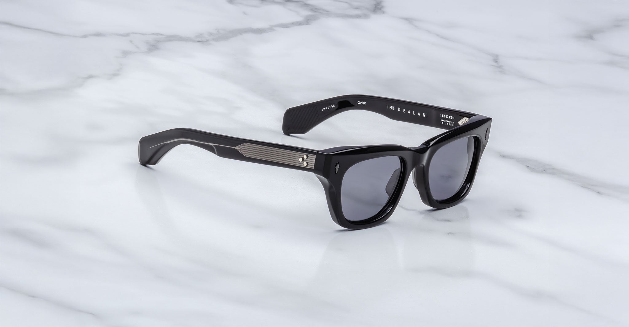 A pair of Jacques Marie Mage Dealan black-framed sunglasses with dark lenses rests on a white marble surface. Silver accents highlight the temples near the hinges.