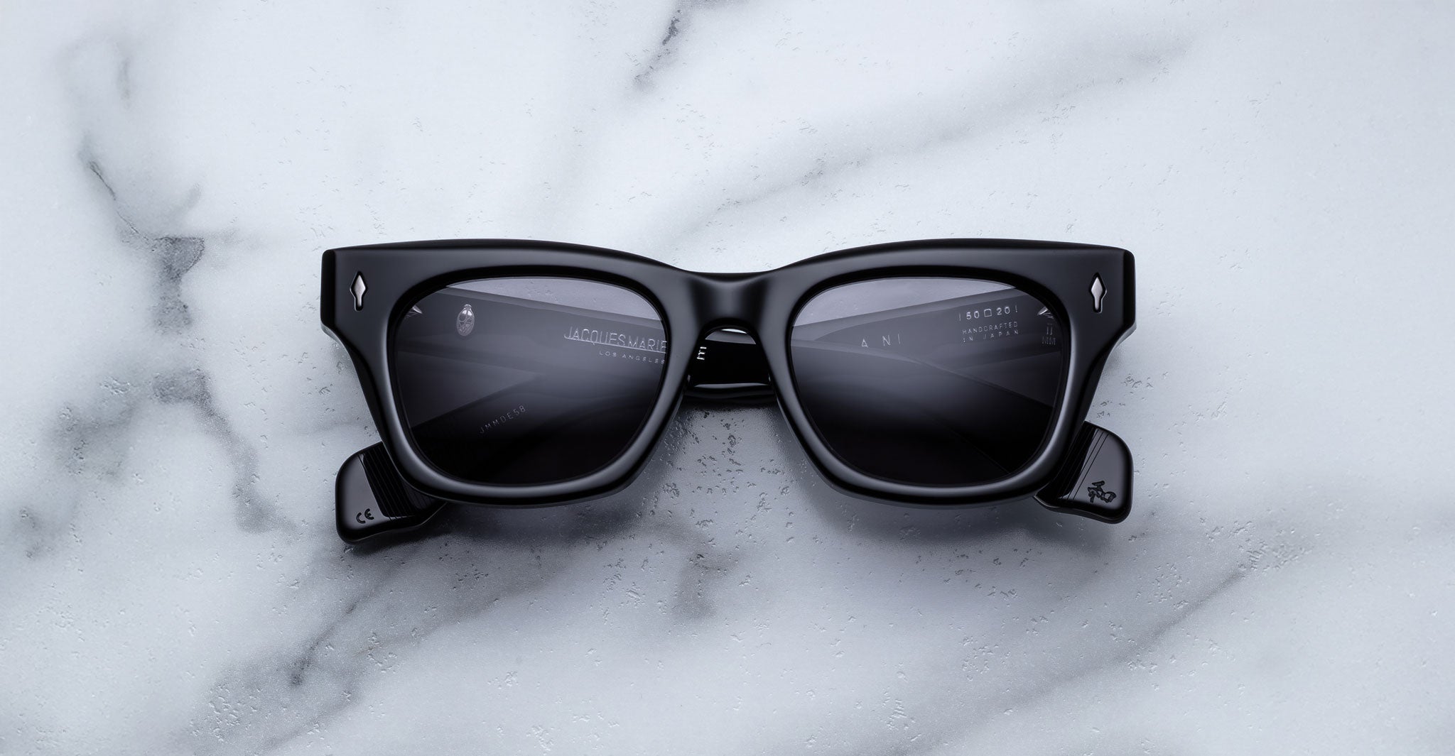 The Jacques Marie Mage Dealan sunglasses in black, featuring slightly reflective dark lenses, rest on a white marble surface with faint digital interface elements visible on the lenses.