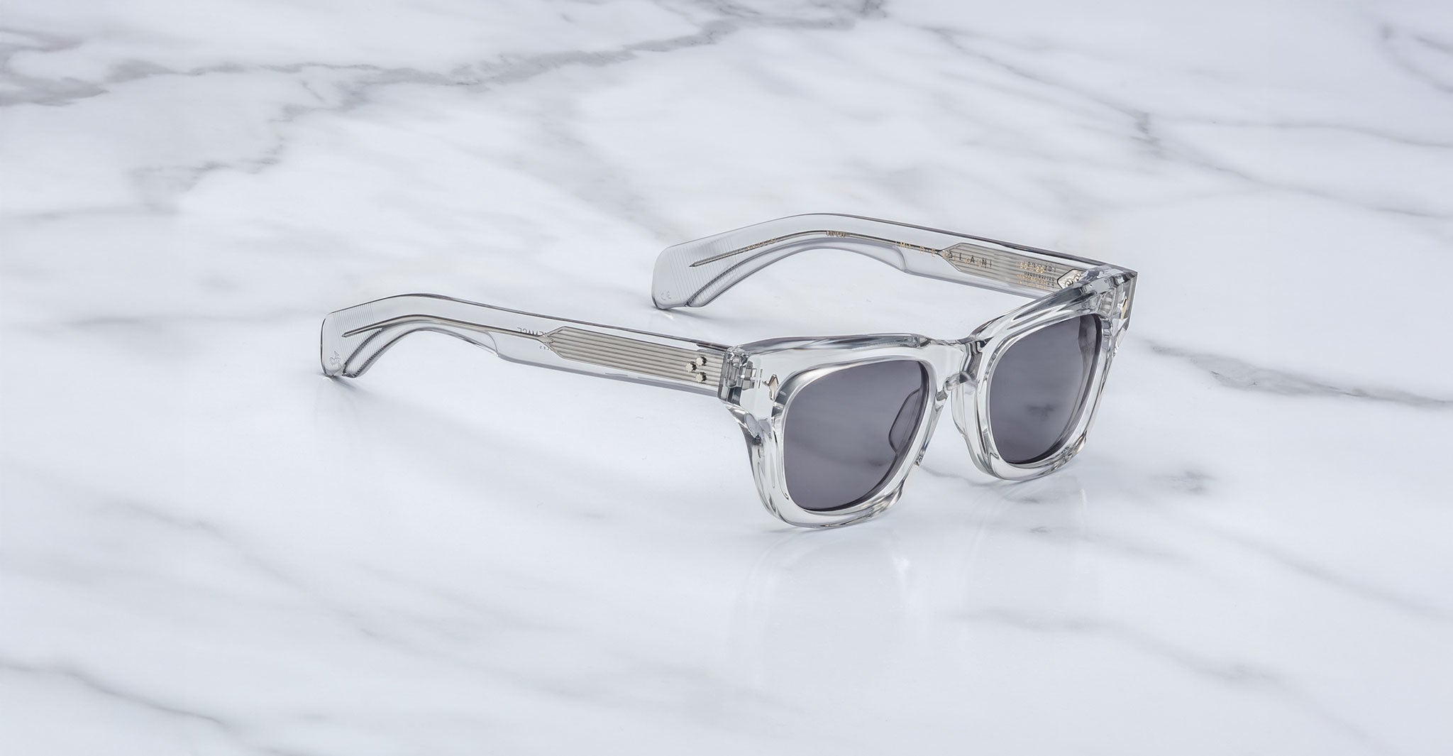 A pair of Jacques Marie Mage Dealan sunglasses with clear frames and dark lenses rests on a white marble surface, featuring subtly striped arms.