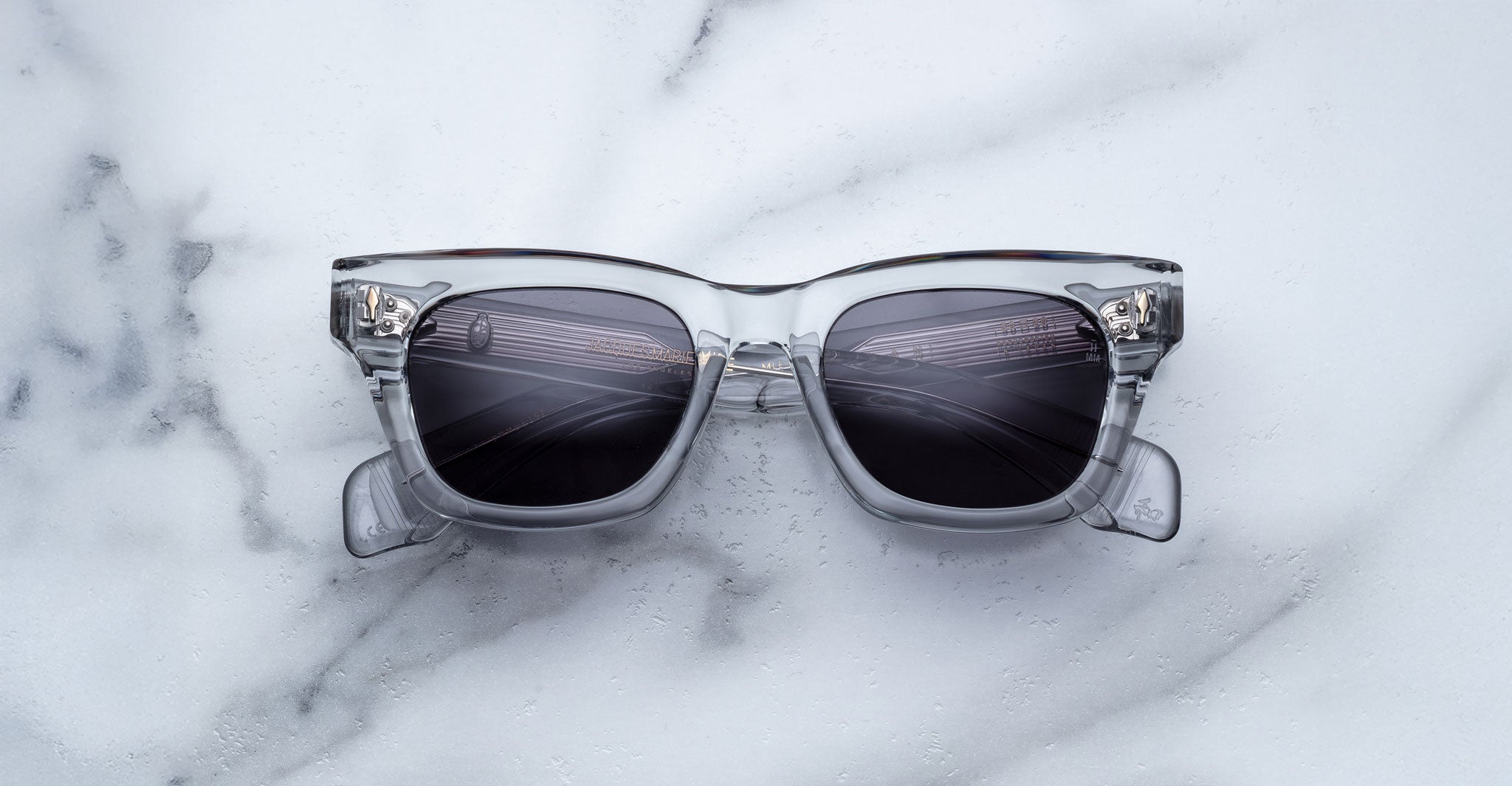 The Jacques Marie Mage Dealan sunglasses, featuring clear squared frames, dark lenses, visible hinges, and transparent arms, are displayed on a white marble surface.