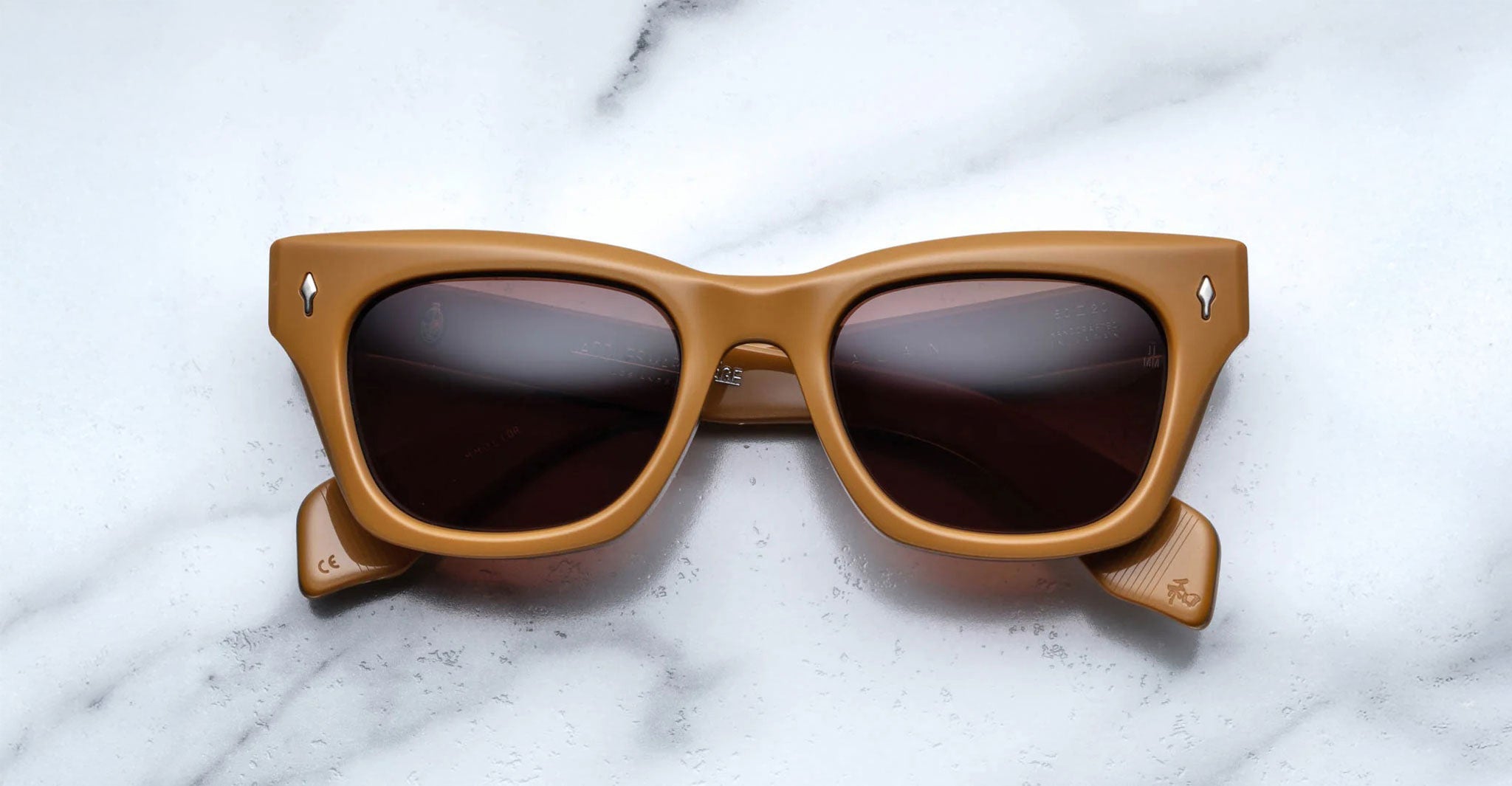 Front view of Jacques Marie Mage Dealan acetate sunglasses in a tan color with dark lenses, sitting on a marbled background