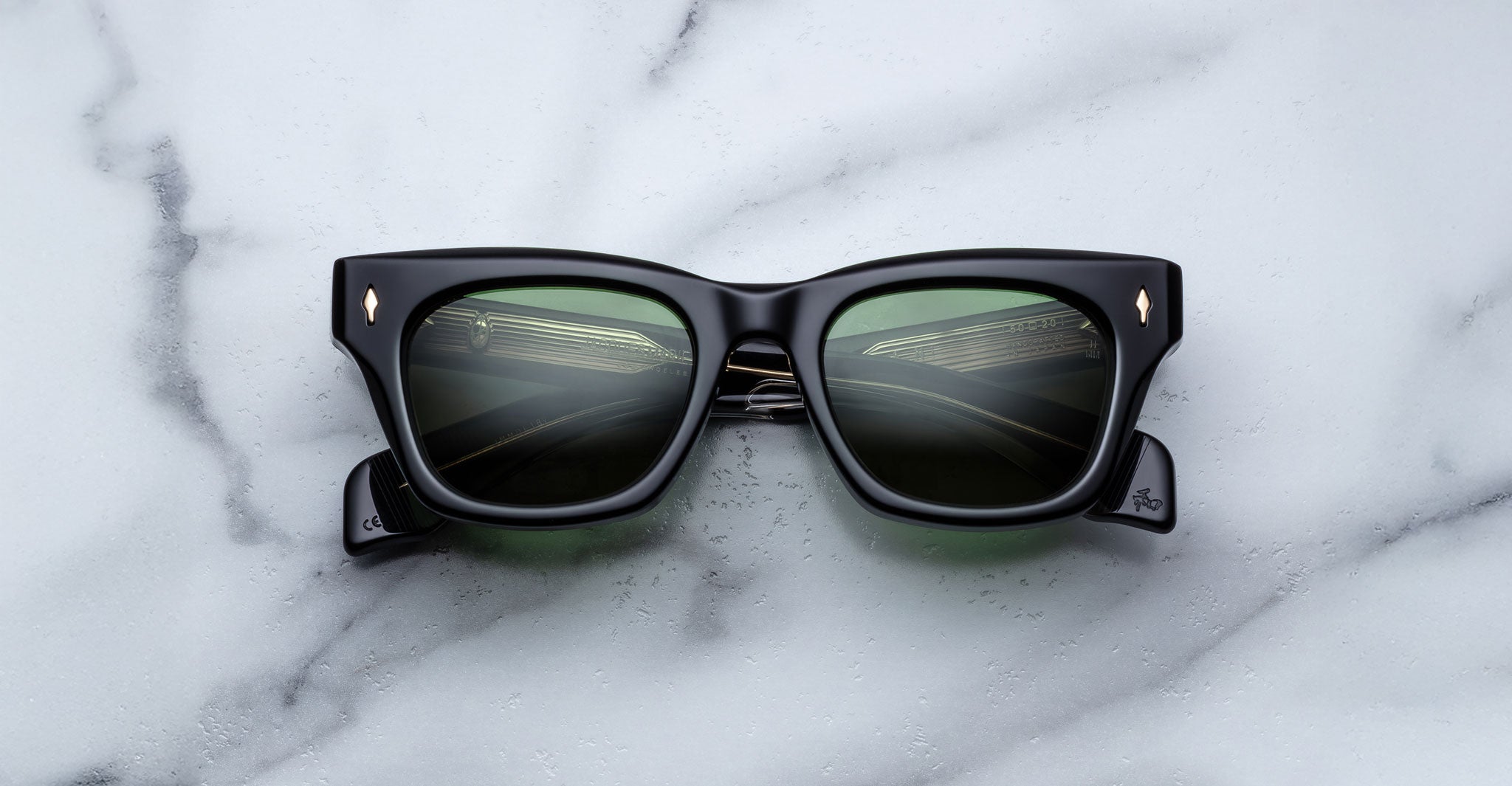 The Jacques Marie Mage Dealan, a pair of black square sunglasses with green-tinted lenses and metal corner accents, rests on a white marble surface.