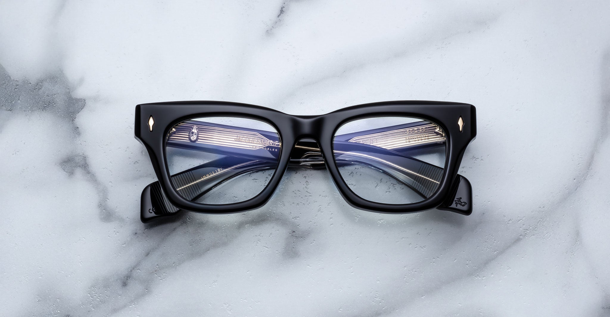 A pair of Jacques Marie Mage Dealan thick black rectangular eyeglasses rests on a white marble surface, with folded temples and lenses reflecting overhead lights.