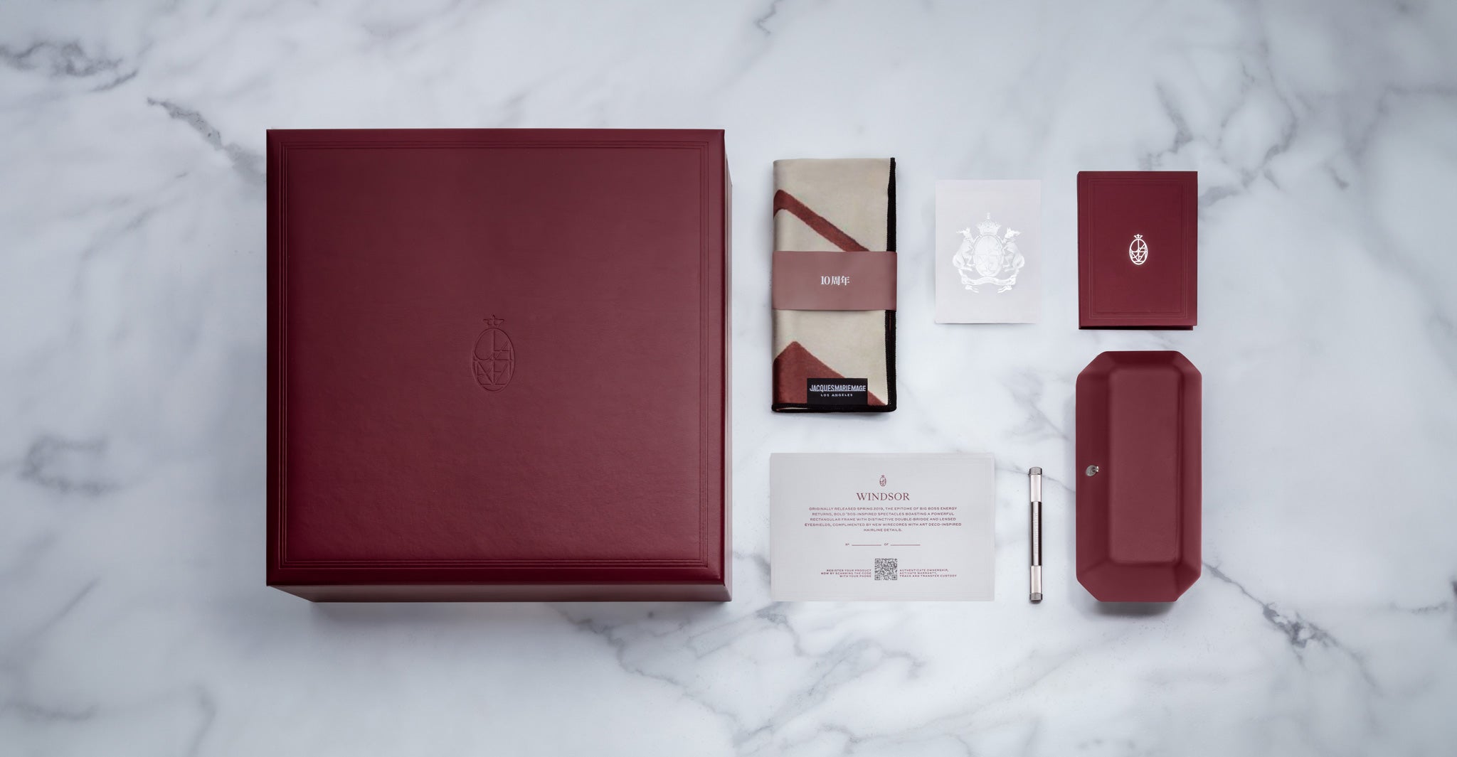 A large burgundy Jacques Marie Mage Windsor box is displayed on a marble surface with matching items: a folded cloth, card, small notebook, rectangular case, pen, and information card.