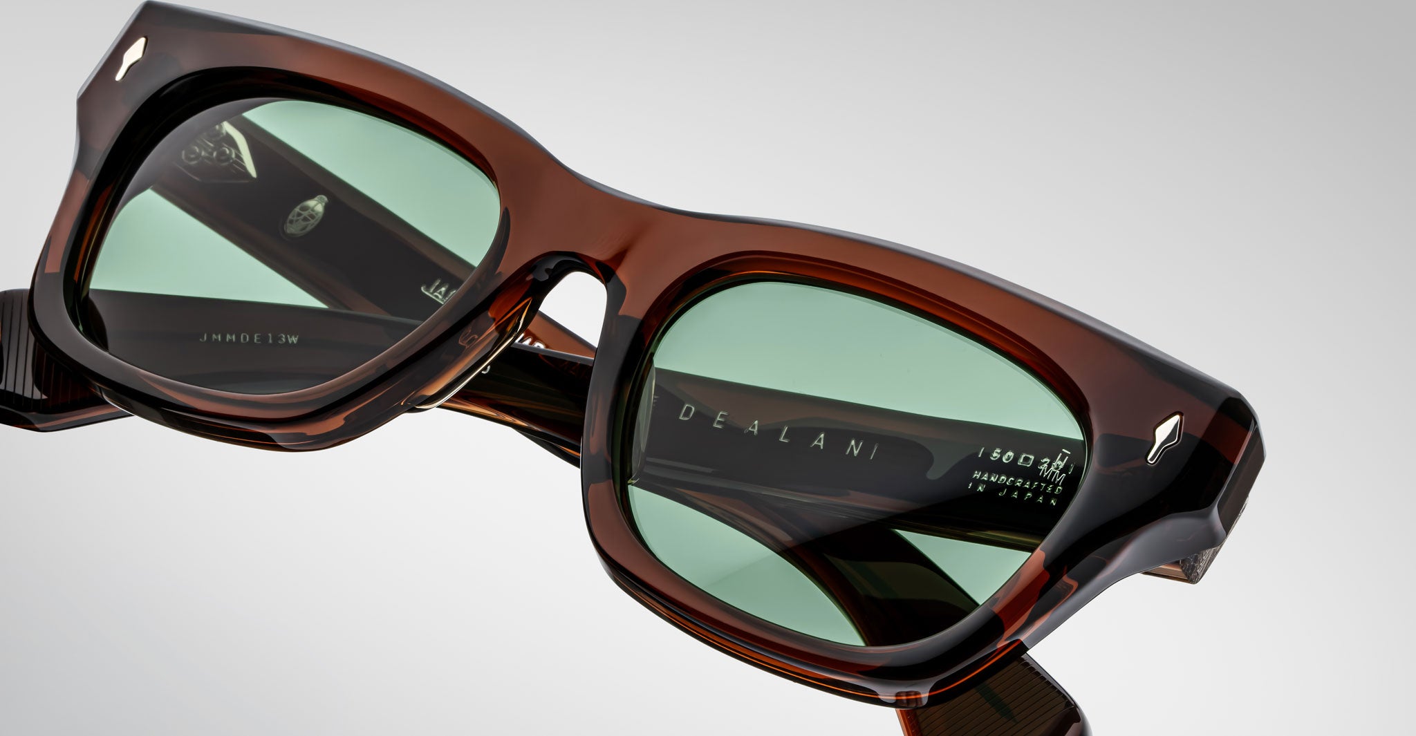 Close-up of Jacques Marie Mage Dealan sunglasses: square, brown acetate frames with green-tinted lenses. The arms feature clear branding and Handcrafted in Japan text, set against a plain light background.