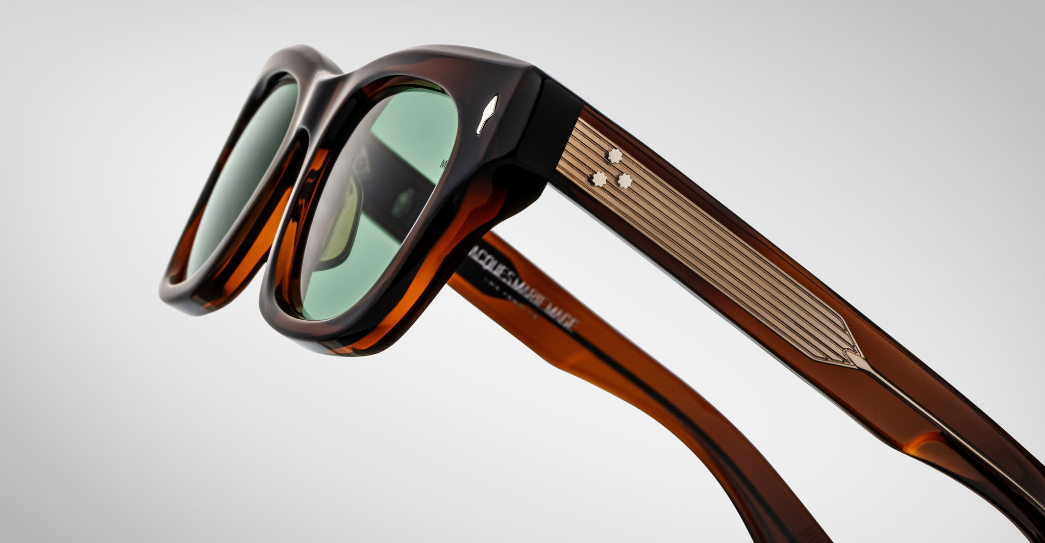 Close-up of Jacques Marie Mage Dealan sunglasses in stylish brown acetate with thick frames, green-tinted lenses, and gold metallic details featuring three decorative dots on the temples against a plain background.